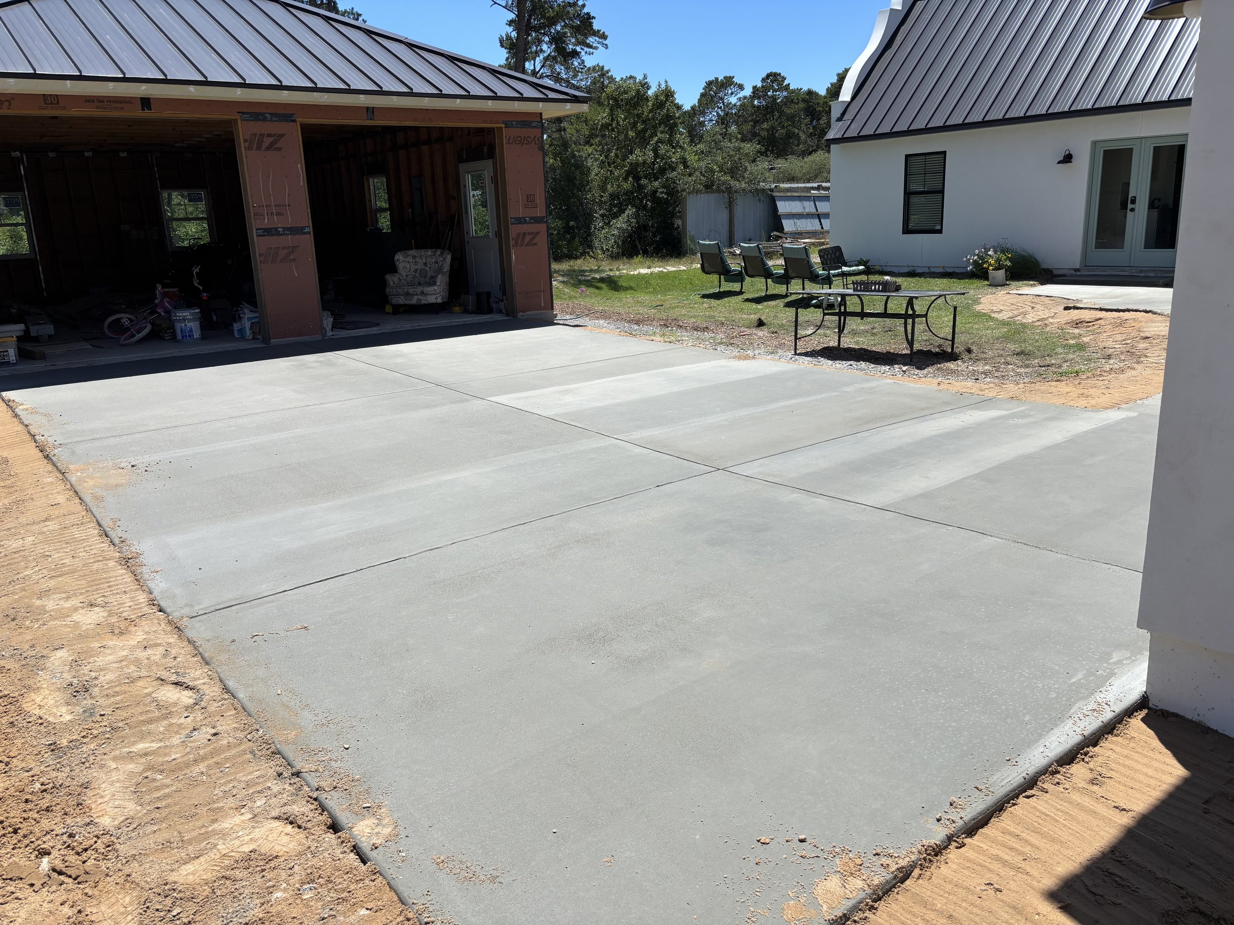 Freshly poured concrete driveway in front of a house under construction, with an unfinished garage to the left and outdoor patio furniture on a grassy area to the right.
