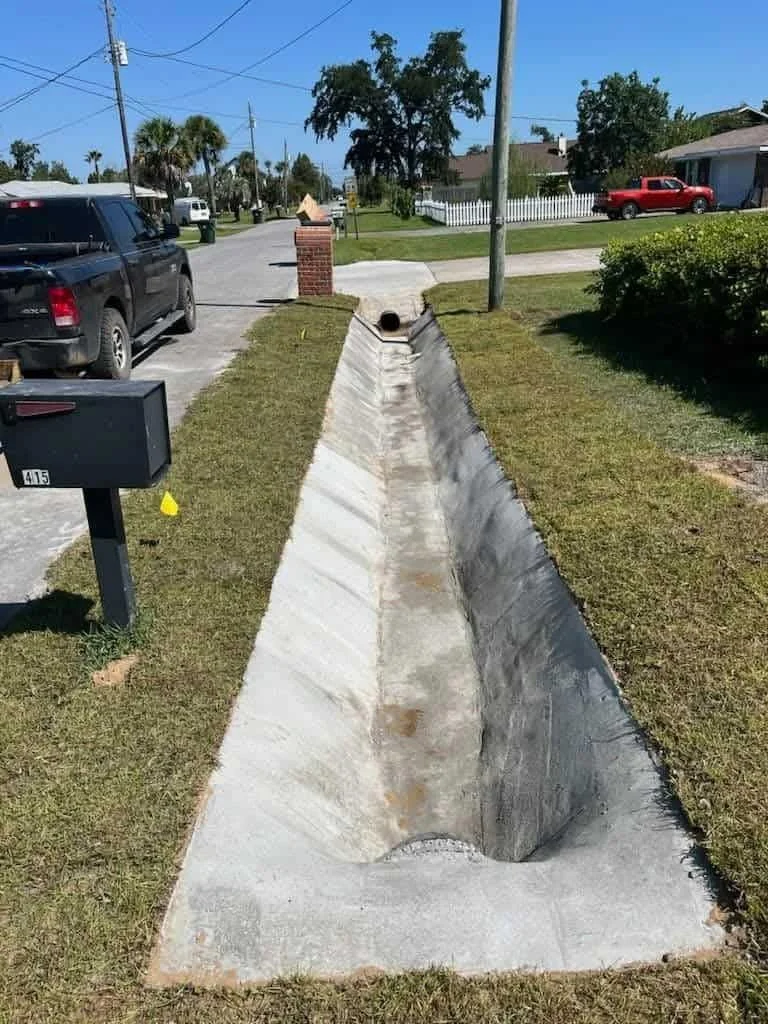 A concrete storm drain trench running through a grass strip in a residential neighborhood on a sunny day.