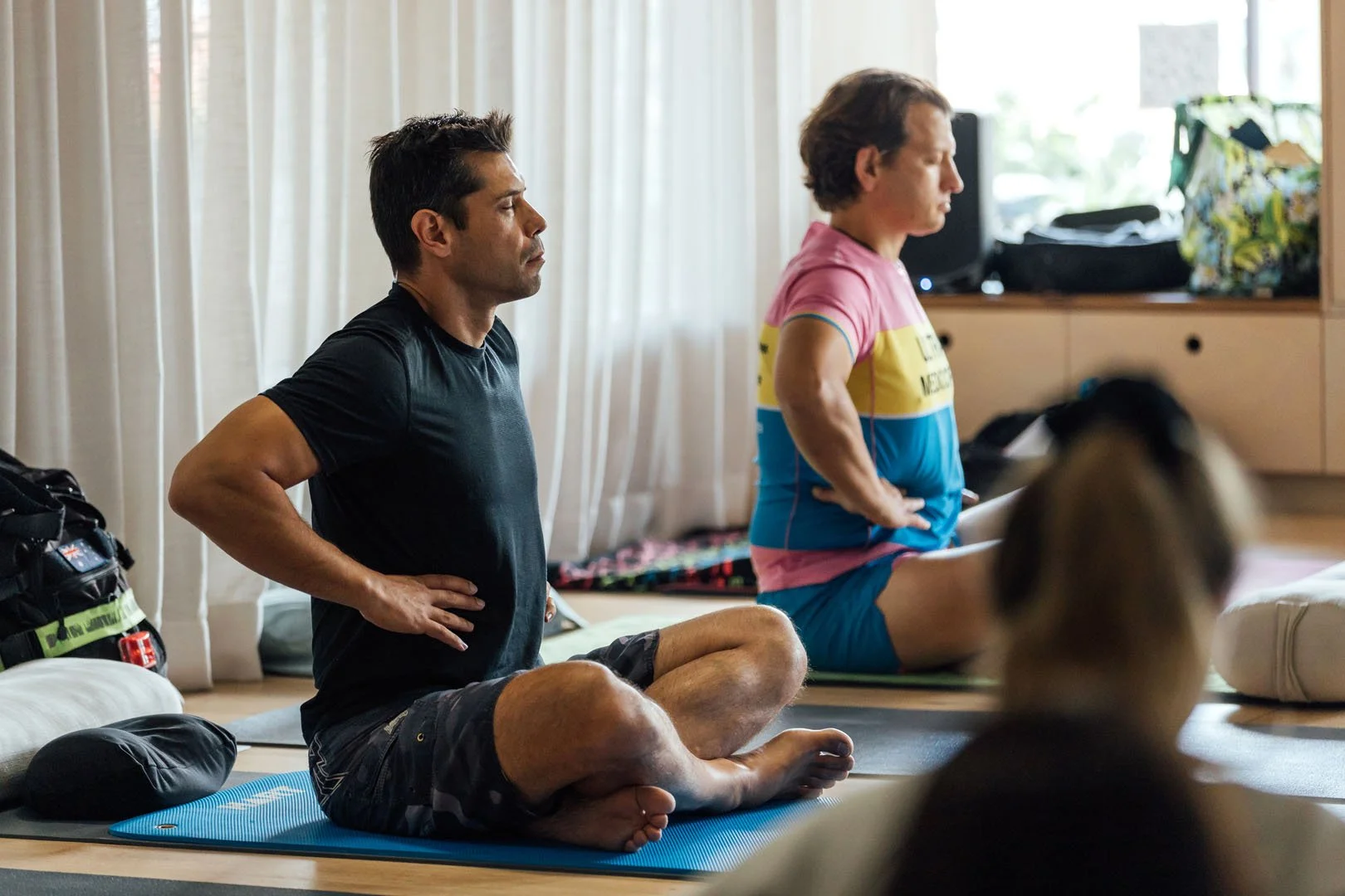 Two men sitting cross-legged on yoga mats with eyes closed, practicing meditation or yoga in a bright room. One man is wearing a black T-shirt and shorts, while the other is in a colorful cycling jersey and shorts.