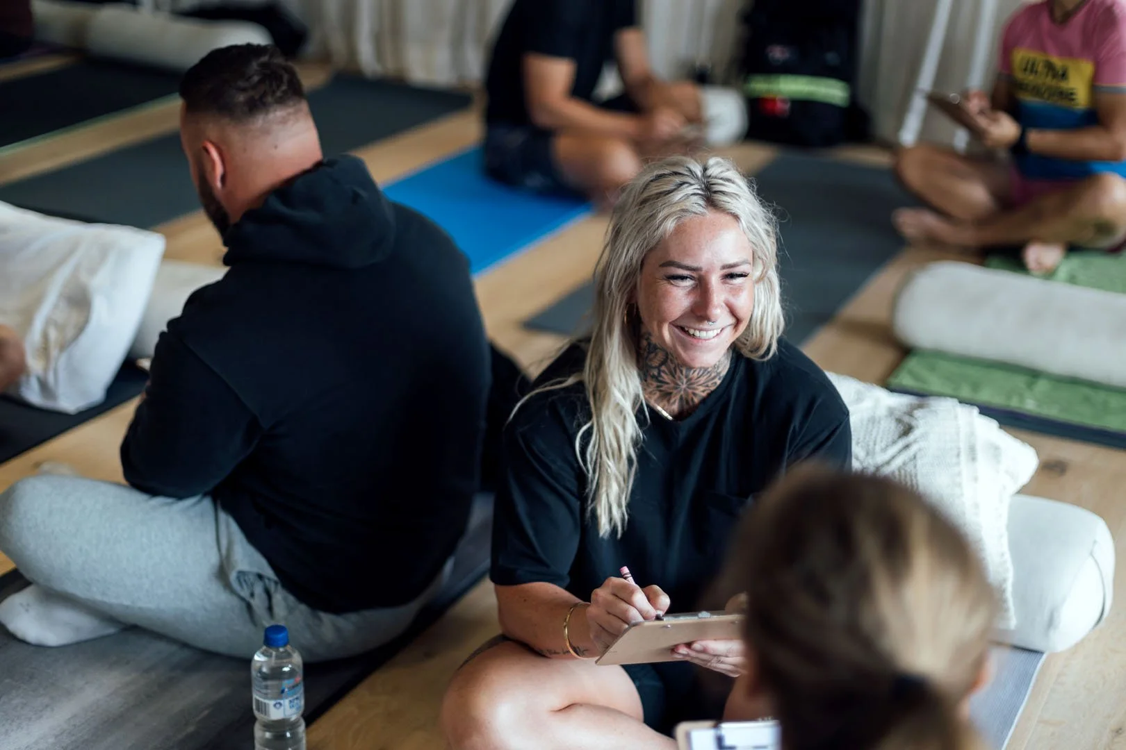 A group of people sitting on yoga mats, with one woman smiling and holding a clipboard, participating in a casual group activity or class in a relaxed indoor setting.