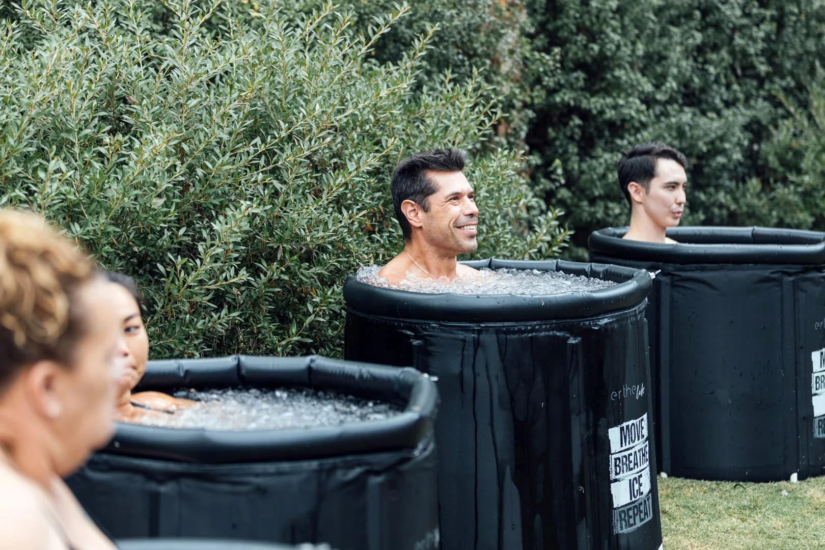 People participating in an outdoor ice bath event, with three individuals submerged in black tubs filled with ice water, green bushes in the background, and one man smiling while relaxing in his tub.