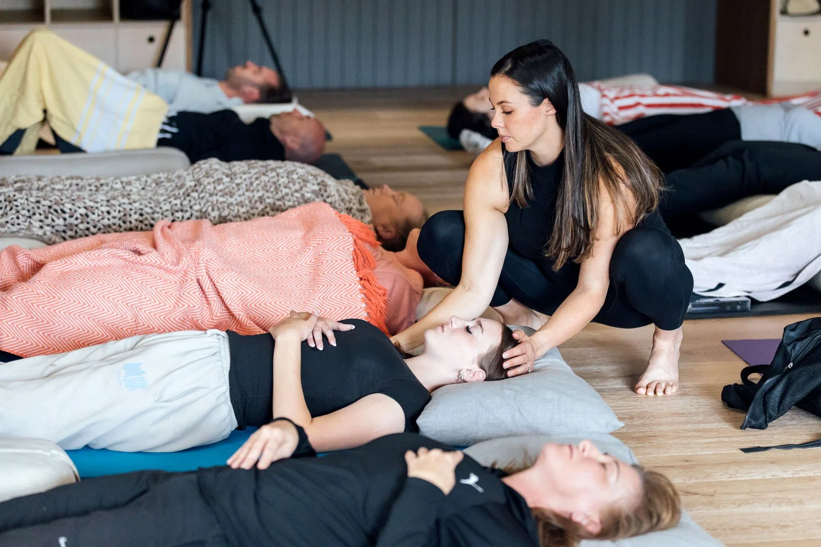Group of people practicing yoga or meditation, lying on the floor with their eyes closed, some with hands on their stomachs or chests, in a calm indoor setting. A woman in black is supporting a woman lying down, gently holding her head and neck.
