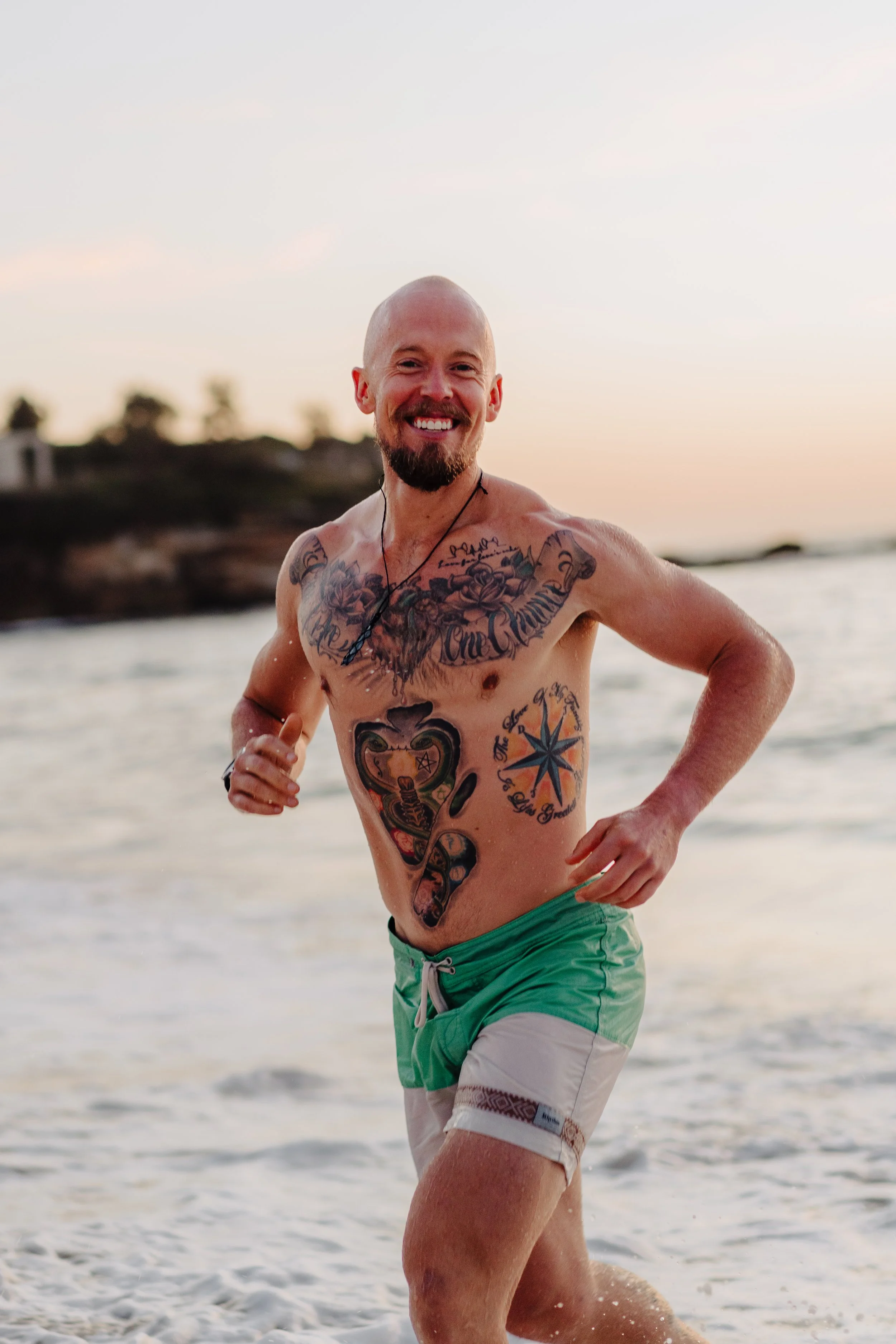 A smiling man with tattoos running in the ocean at sunset, wearing green and beige swim trunks.