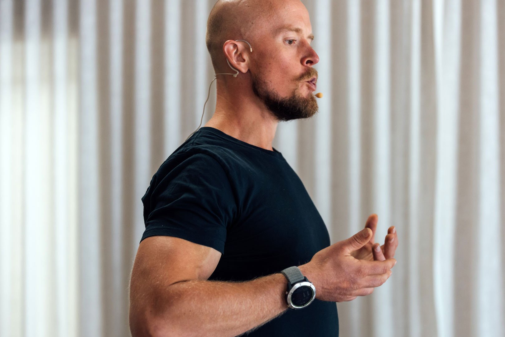 A man with a beard and bald head speaking, wearing a black t-shirt, a wristwatch, and an earpiece, with curtains in the background.