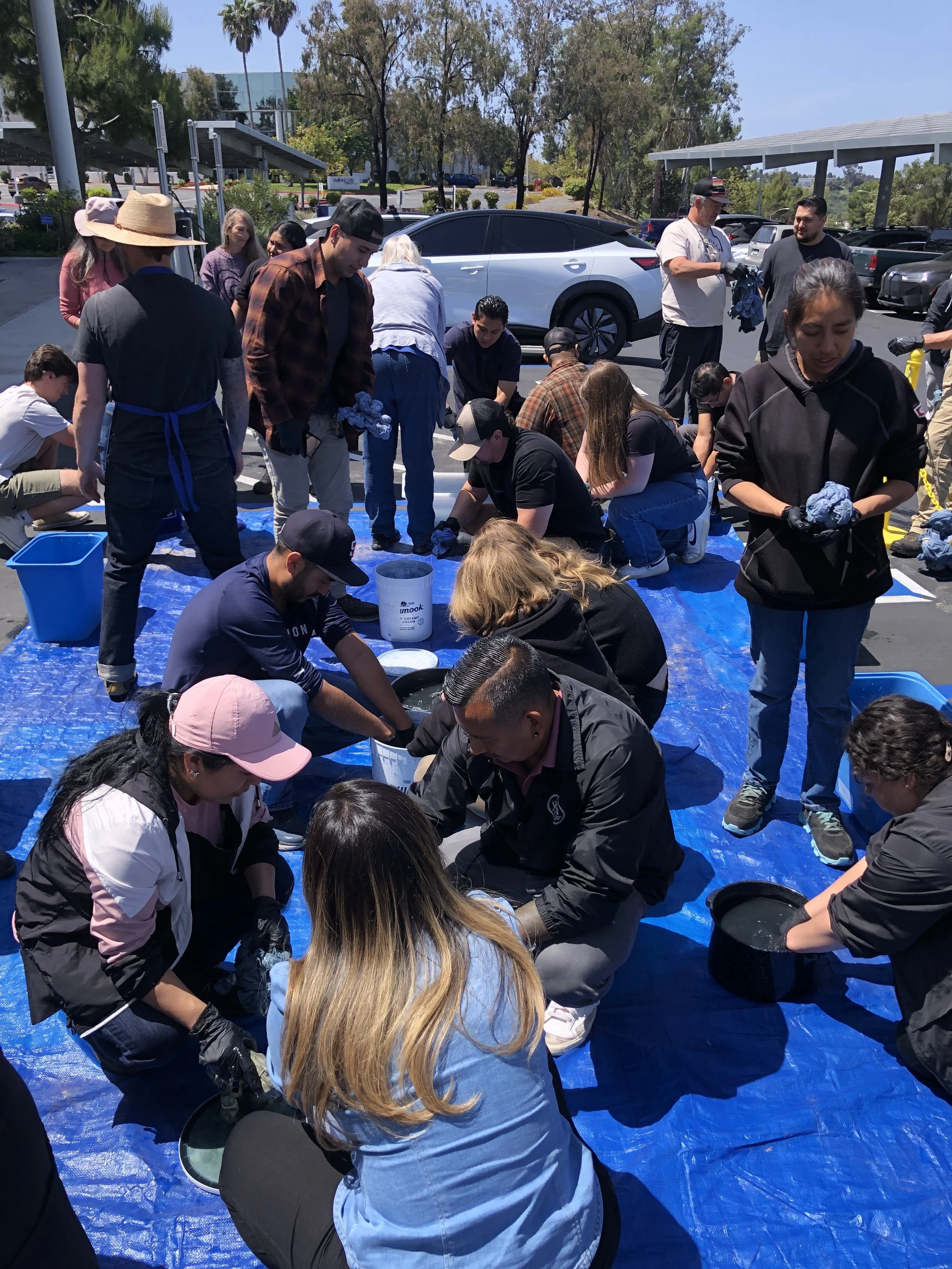 A group of people gathered around and observing a large tank of a liquid placed on a blue tarp in a parking lot, with some individuals wearing gloves and holding cloths or tools.