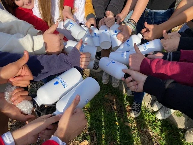 Group of kids outdoors holding white plastic bottles with blue labels, showing thumbs up.