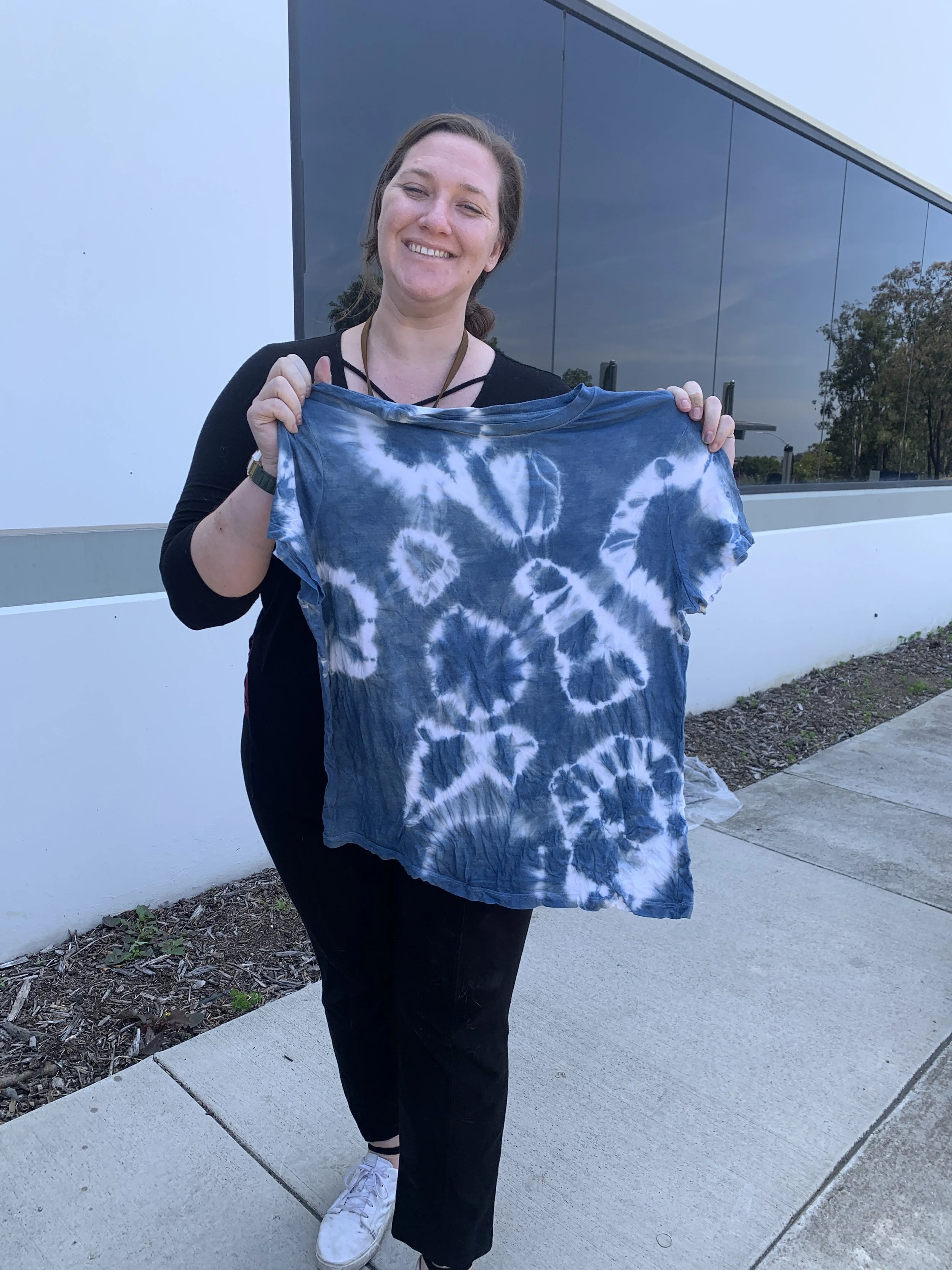 A woman smiling and holding a tie-dye t-shirt outside near a modern building with reflective glass windows.