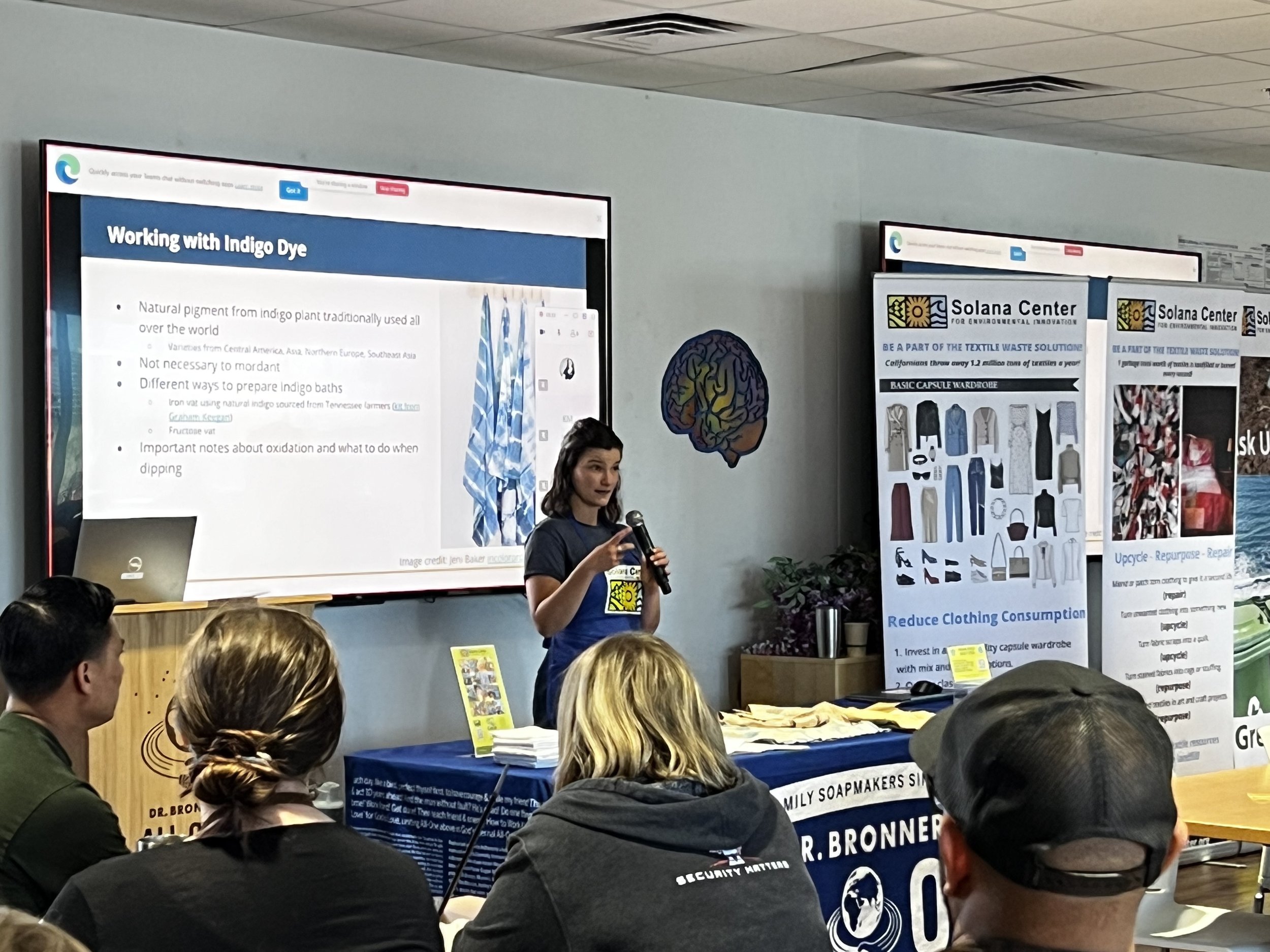 A woman is giving a presentation about working with indigo dye at a conference. She is holding a microphone and standing in front of a screen displaying information about indigo dye. Several audience members are seated, listening attentively.