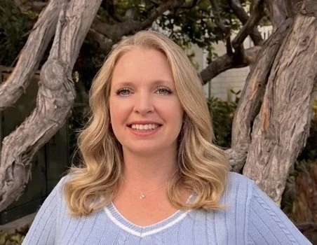 Woman with blonde hair smiling outdoors in front of twisted tree branches, wearing a light blue sweater.