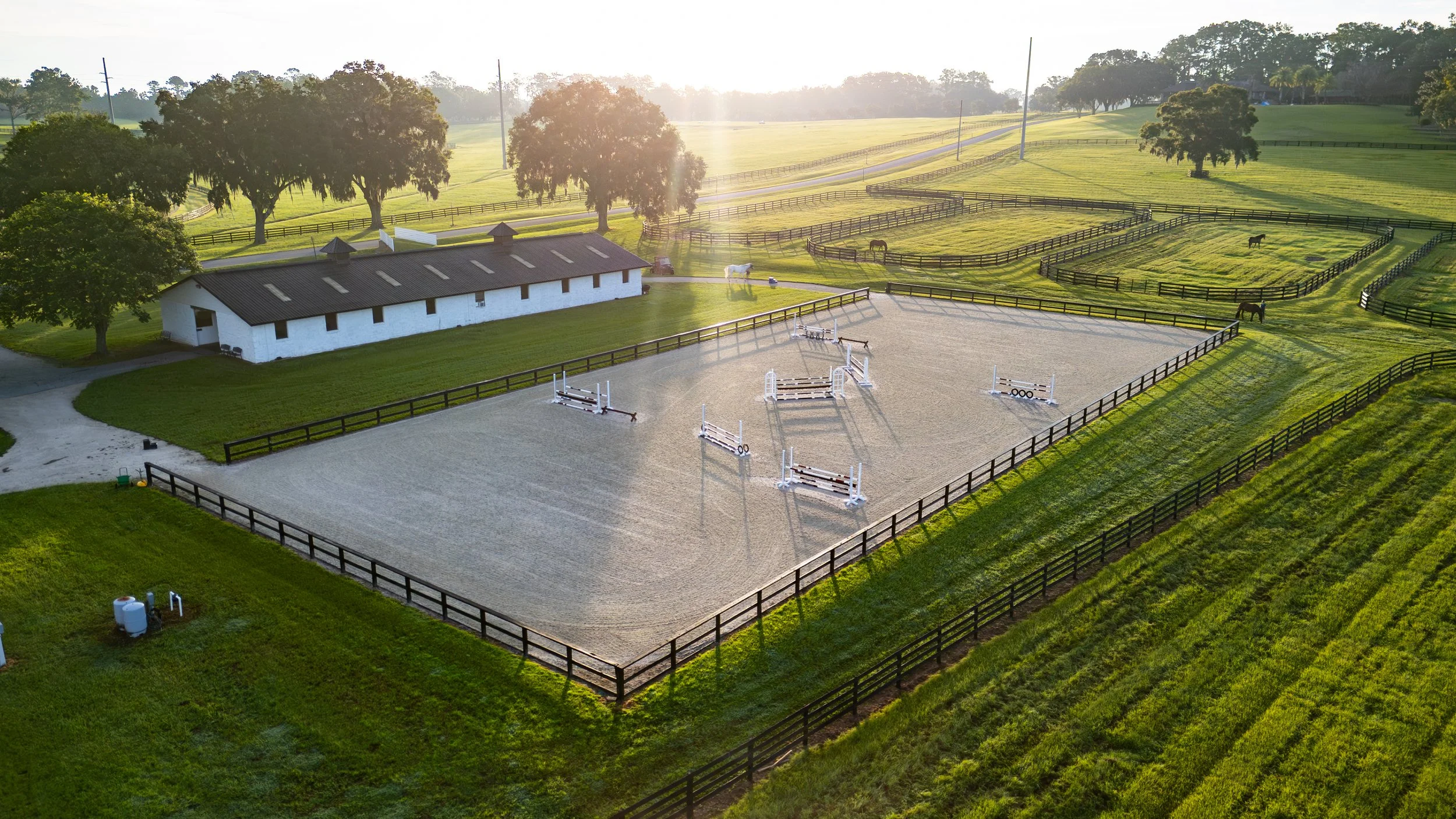 An outdoor horse riding arena with several jumps, surrounded by a black wooden fence, with green grassy fields, trees, and distant hills under a setting sun.