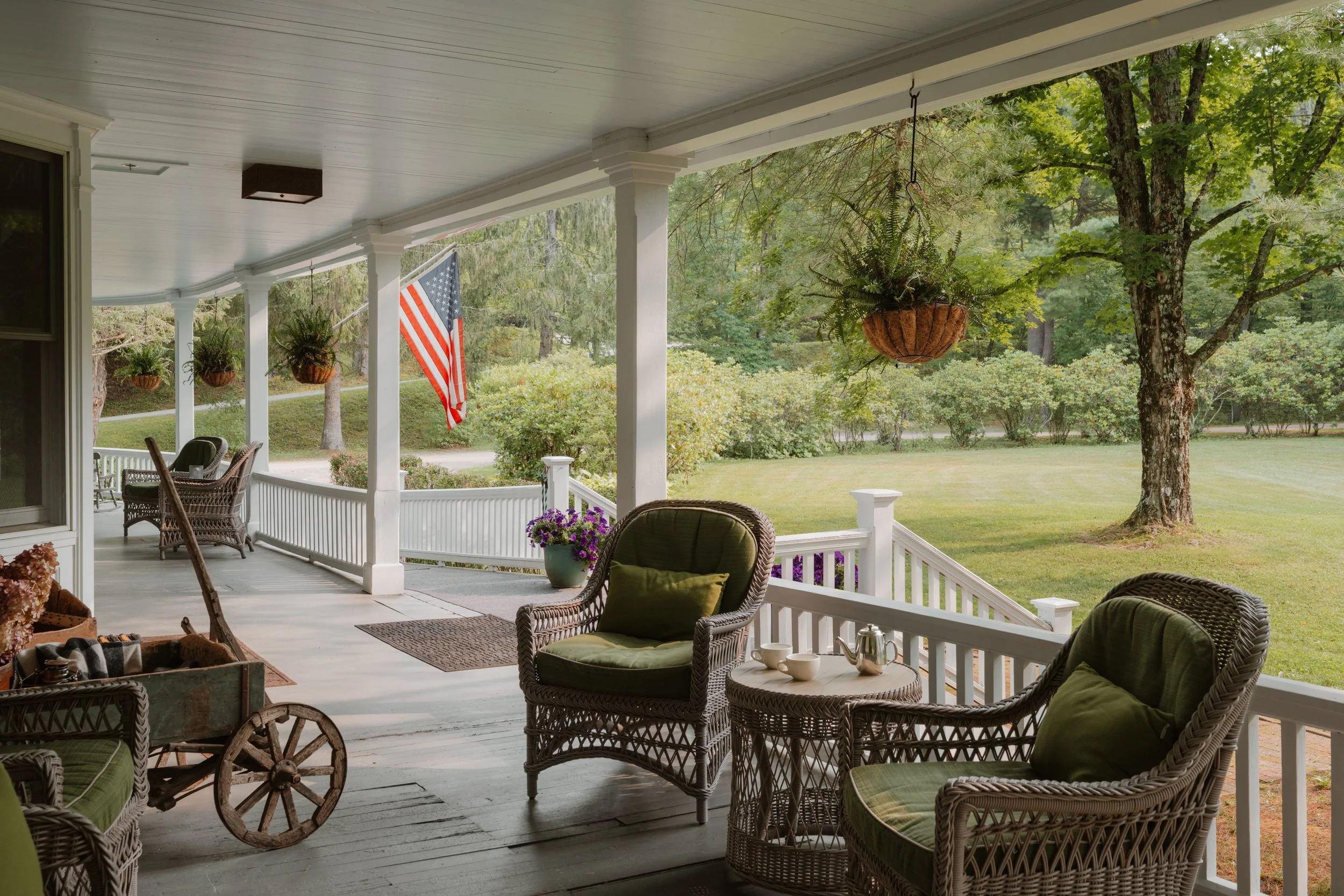 Covered porch with wicker chairs, green cushions, a small table with teapot and cups, hanging plants, potted flowers, American flag, and a scenic view of a lawn with trees.