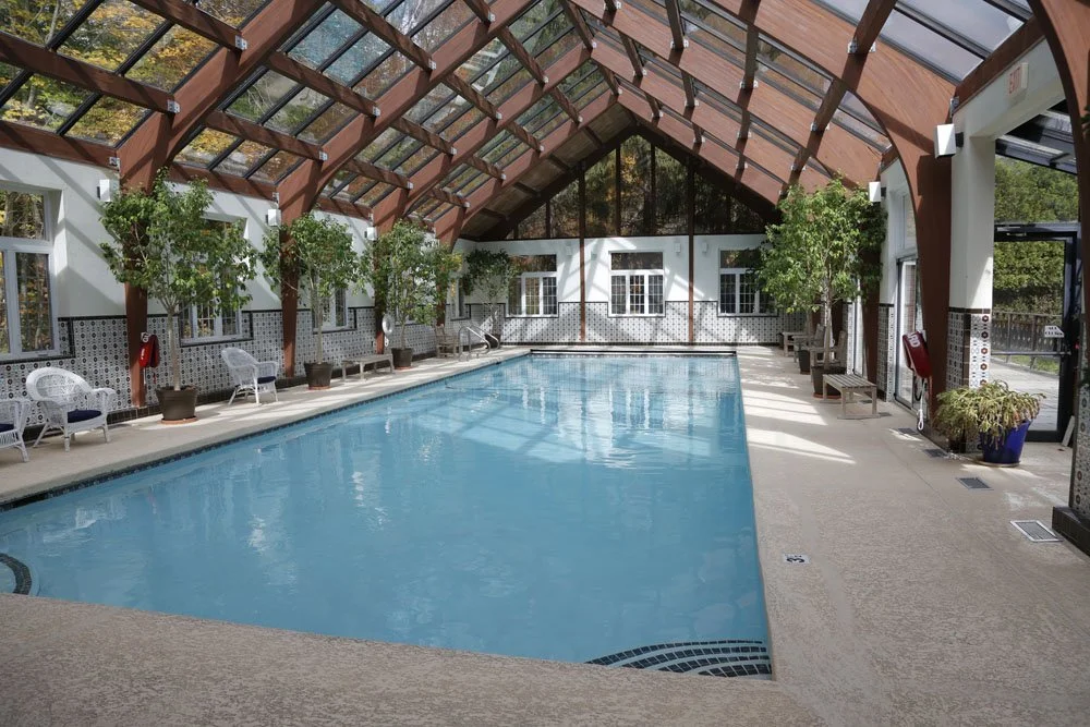 Indoor swimming pool area with a glass vaulted ceiling, white walls, potted trees, and lounge chairs along the sides.