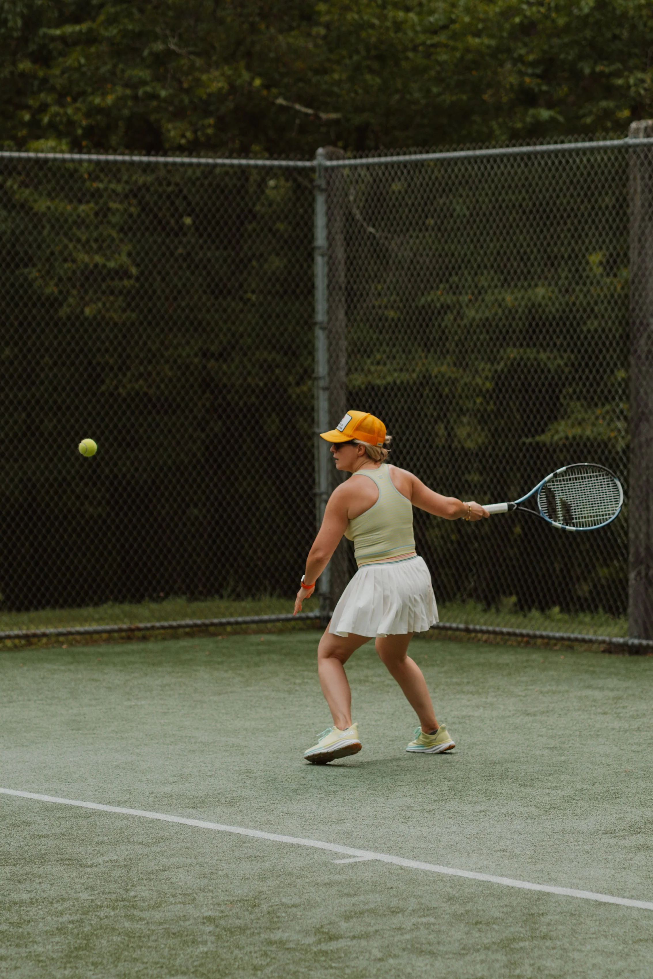 A woman in a green tank top, white tennis skirt, and orange cap playing tennis on an outdoor court with trees in the background.