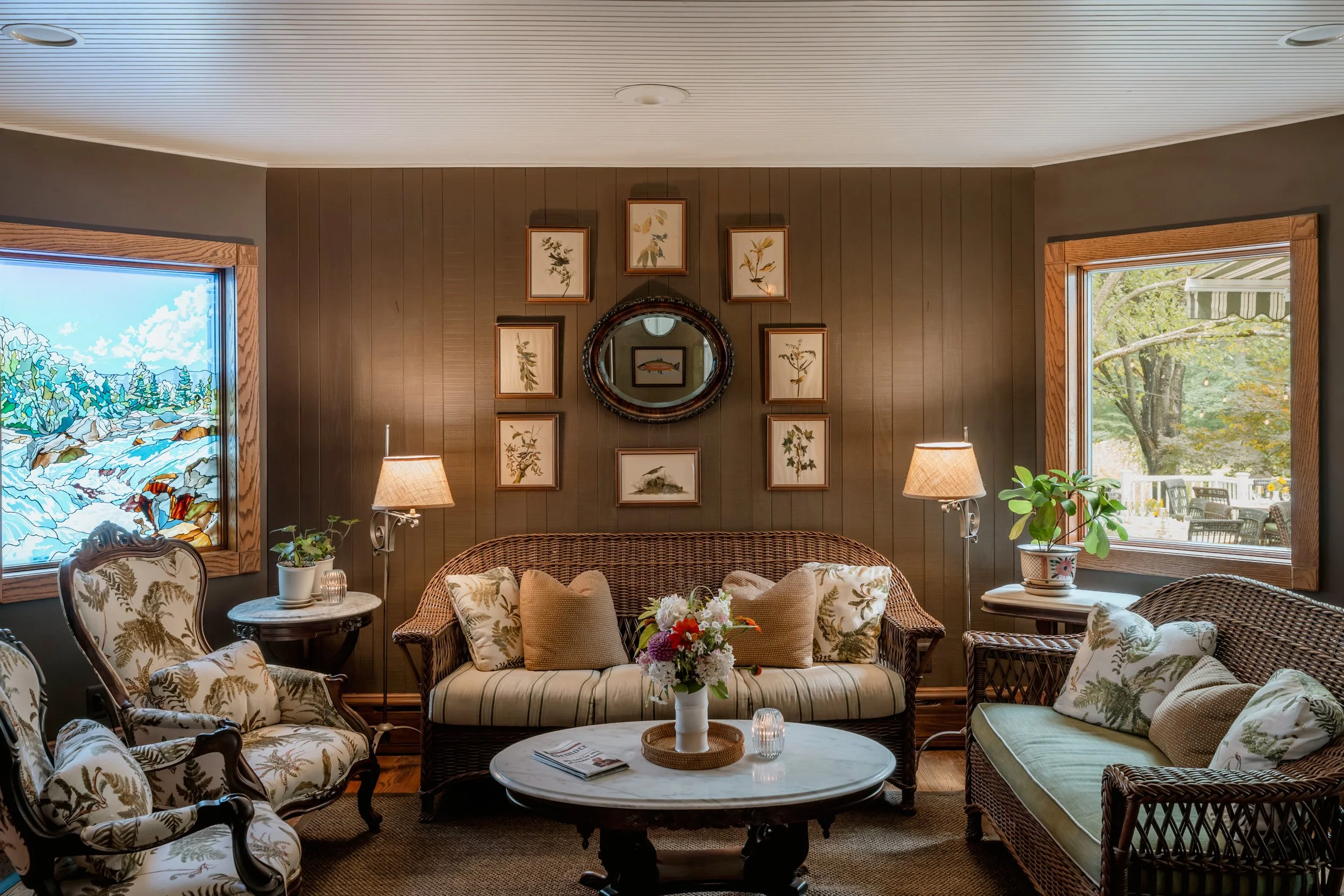 Cozy living room with wooden-paneled walls, floral-patterned furniture, a marble-topped coffee table with flowers, and two large windows showing outdoor greenery.