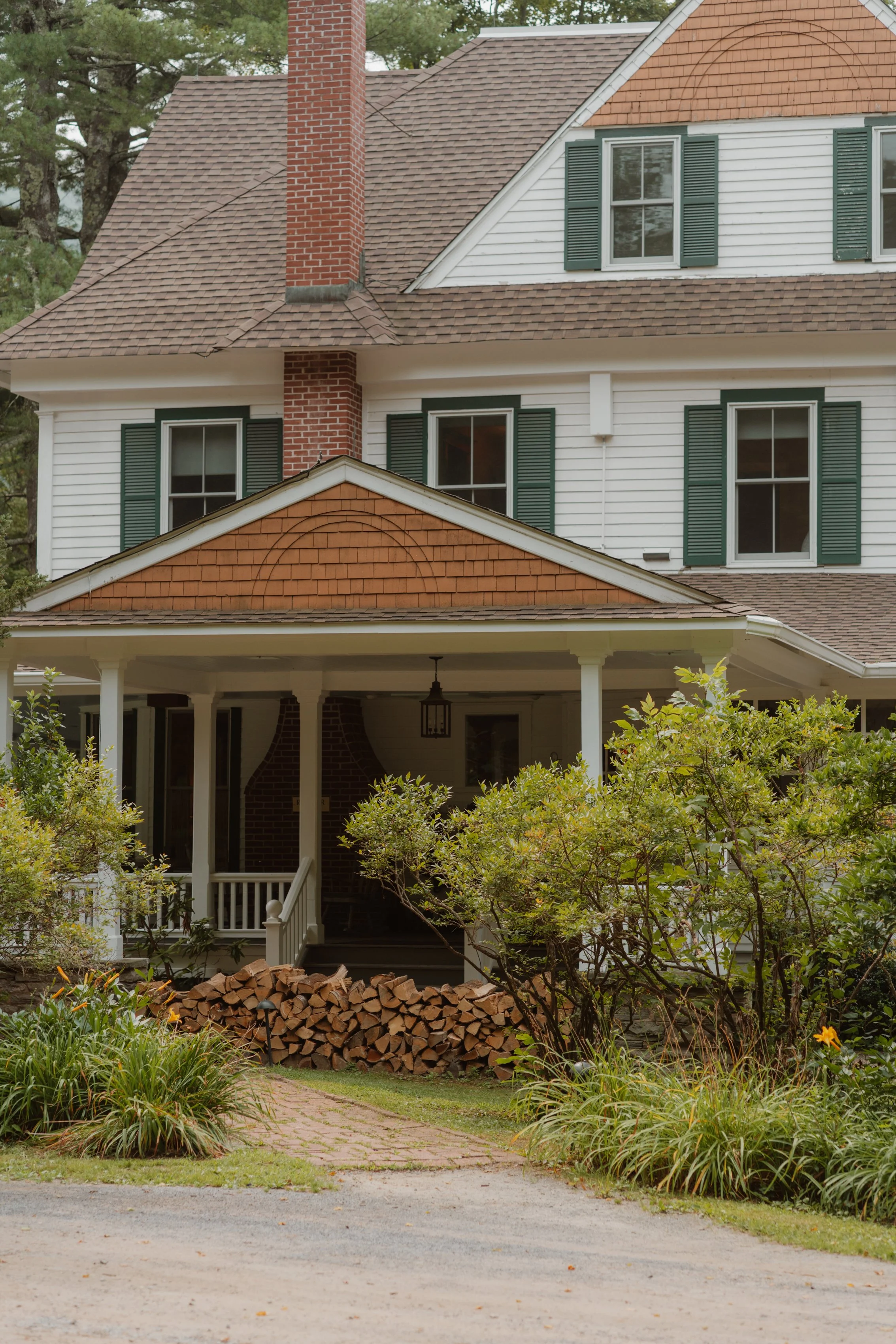 A multi-story house with white exterior siding, green shutters, and a brick chimney. The house has a front porch with a pile of chopped firewood and is surrounded by greenery and landscaping.