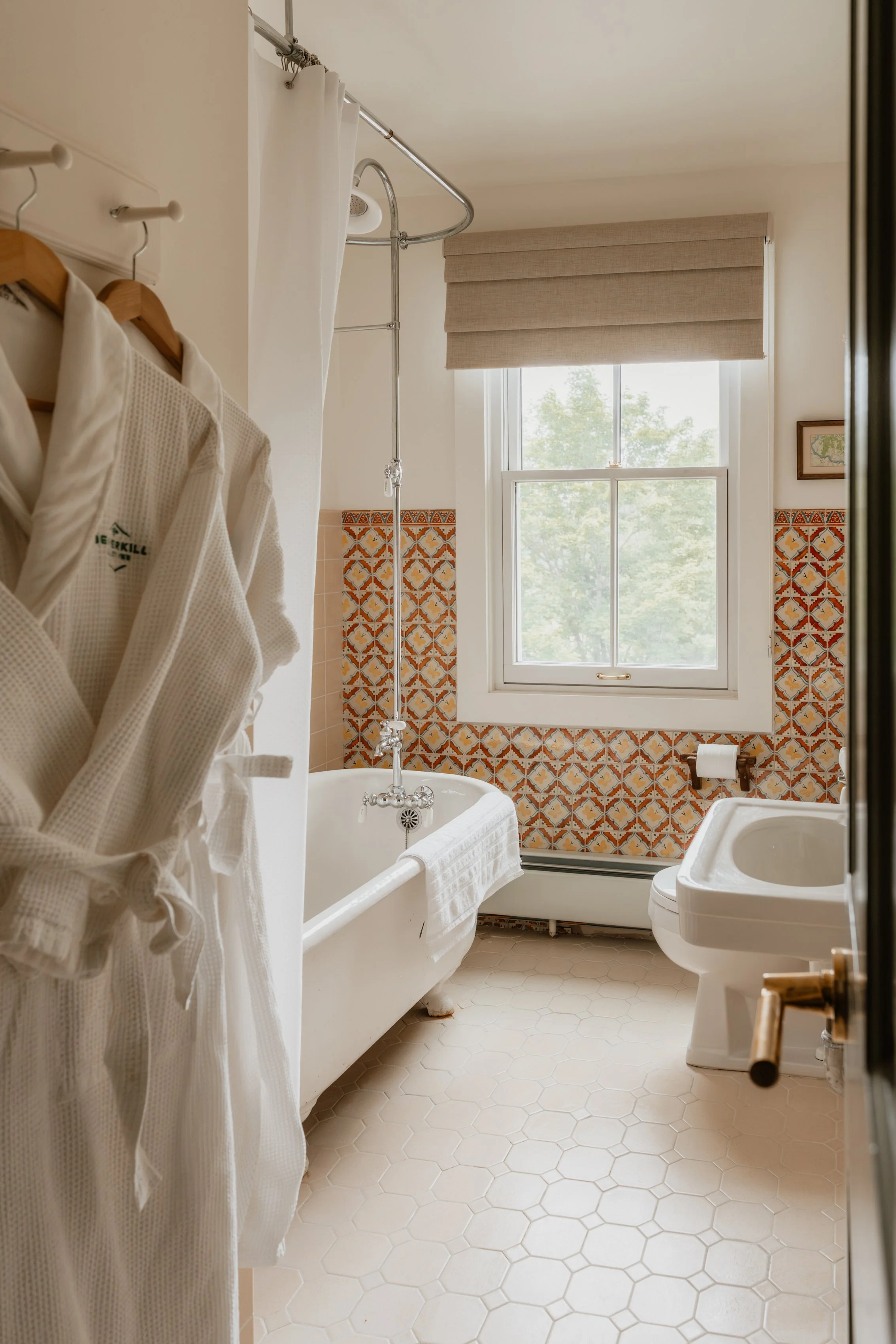 Bathroom with a white bathtub, wooden hangers with bathrobes, a window with beige blinds, and vintage tile decor.