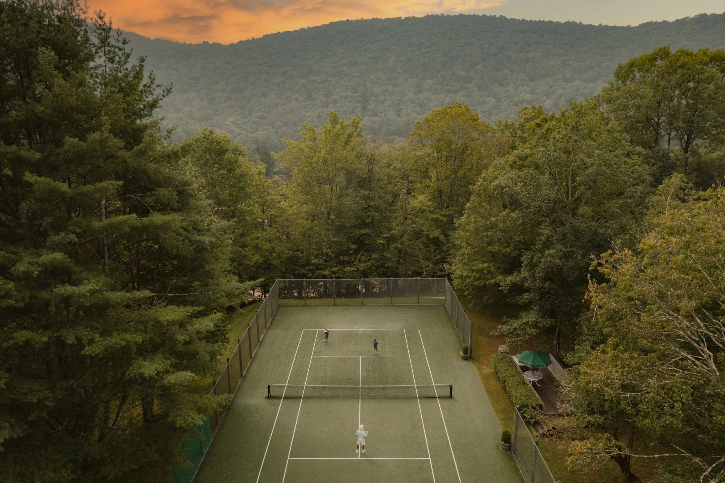 A tennis court surrounded by trees and mountains at sunset with three people playing tennis.