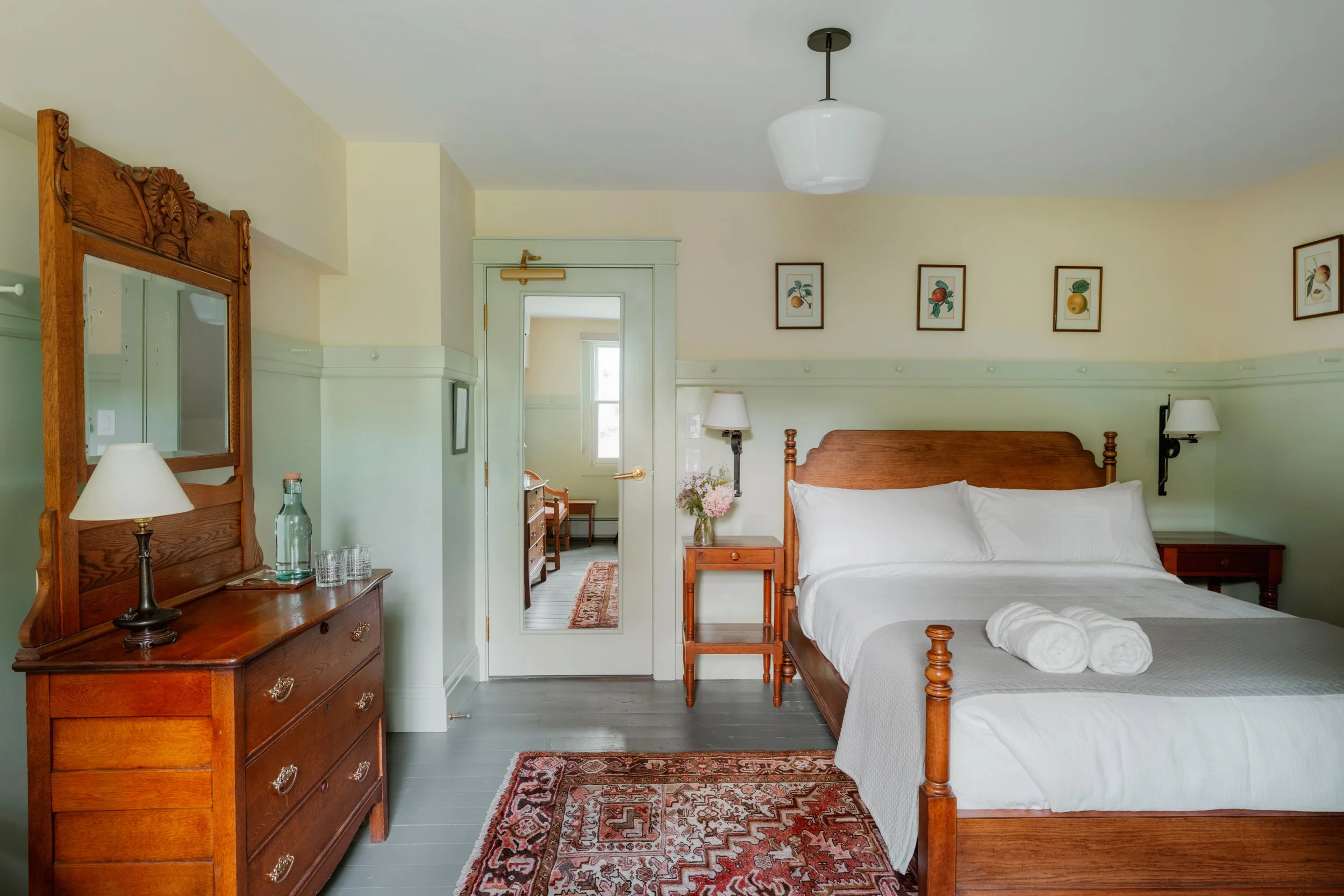 A cozy bedroom with a wooden bed, matching nightstands, and a dresser with a mirror. The room has light-colored walls, framed fruit-themed artwork, and a patterned rug. Towels are neatly rolled on the bed, and there's a vase of flowers on the nightst