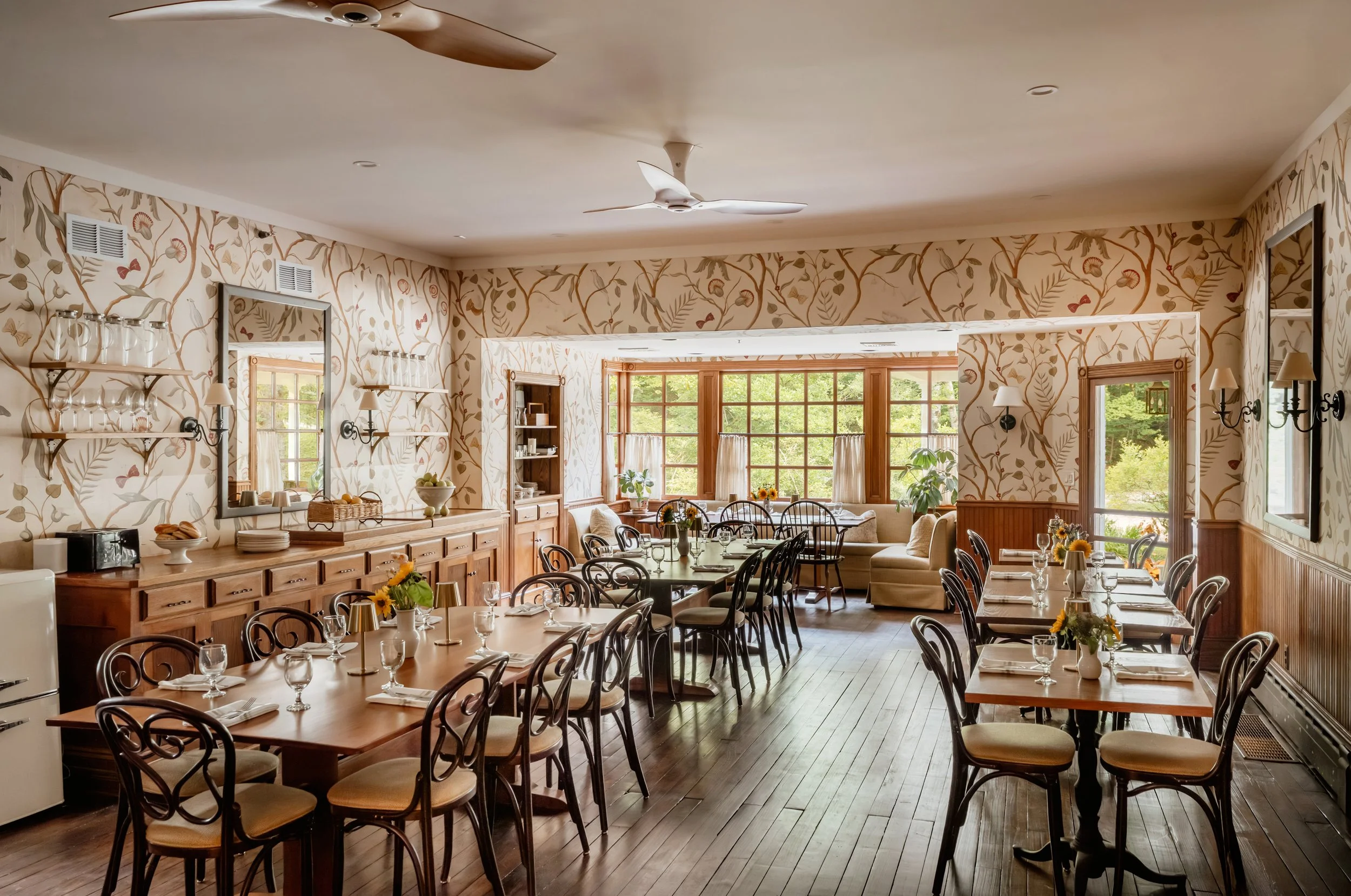 A dining room with wooden tables set with wine glasses, napkins, and small flower arrangements, decorated with floral wallpaper, wooden wainscoting, and large windows letting in natural light.