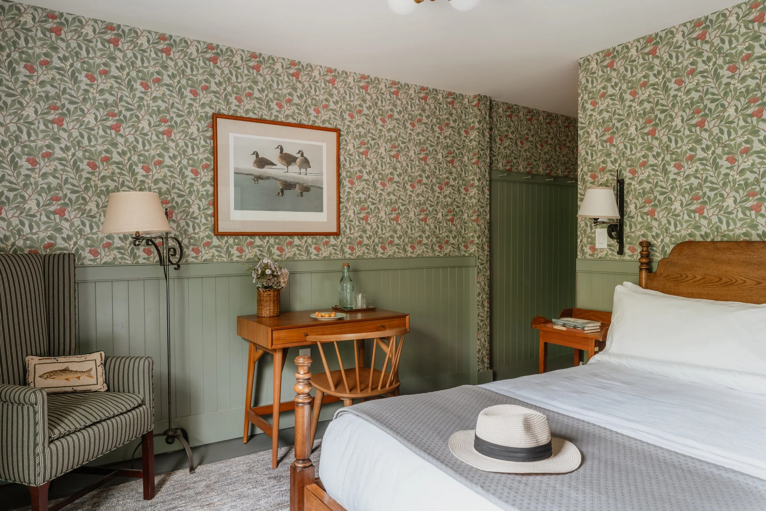 Cozy bedroom with green floral wallpaper, a wooden bed with white sheets and a straw hat, a wicker chair, a small wooden desk with a vase and water bottle, and framed bird artwork on the wall.