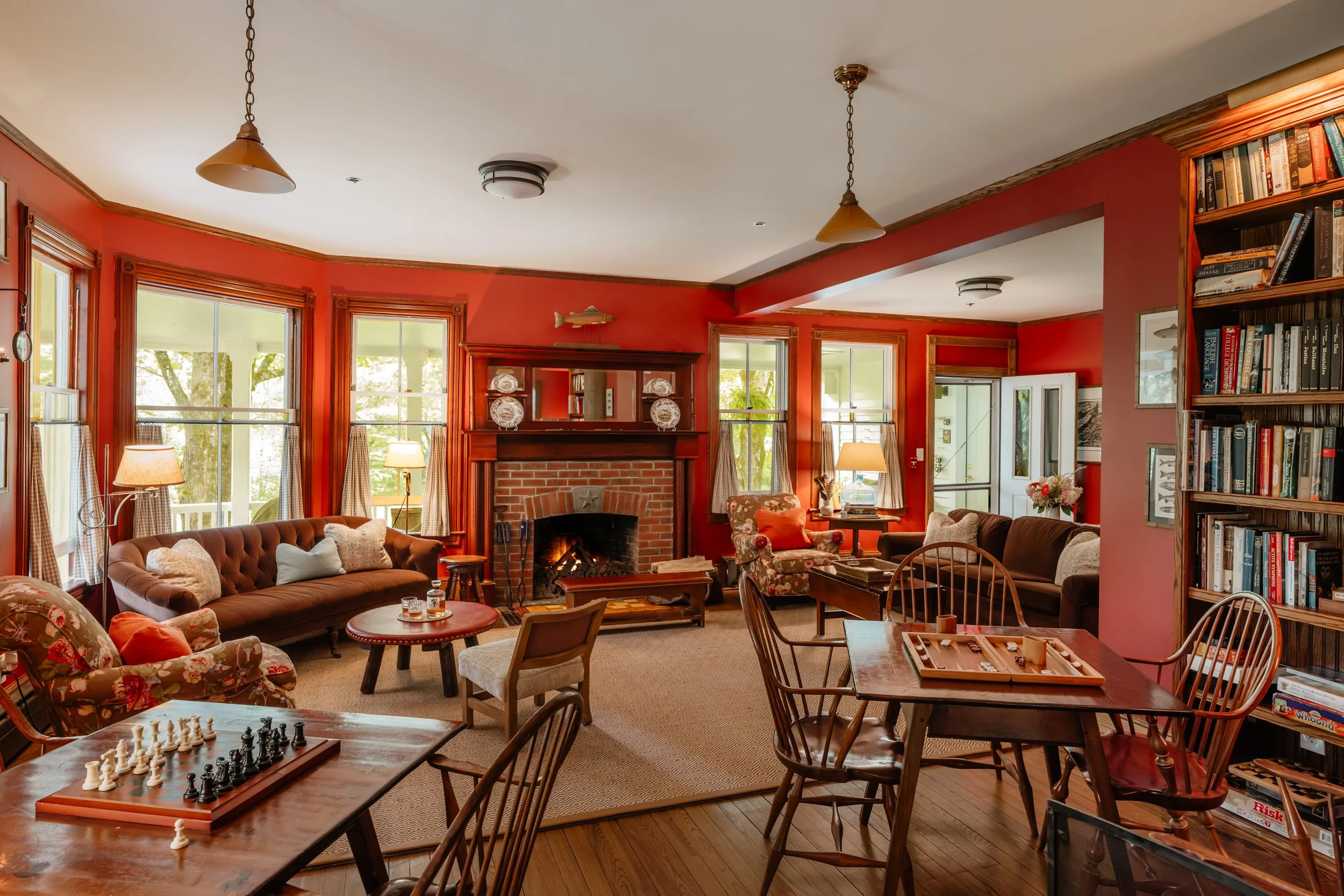 Cozy living room with red walls, multiple windows, a brick fireplace, and various seating options surrounding a central coffee table. Several chairs are around small tables, with a chess set on one. There are bookshelves filled with books and decorat