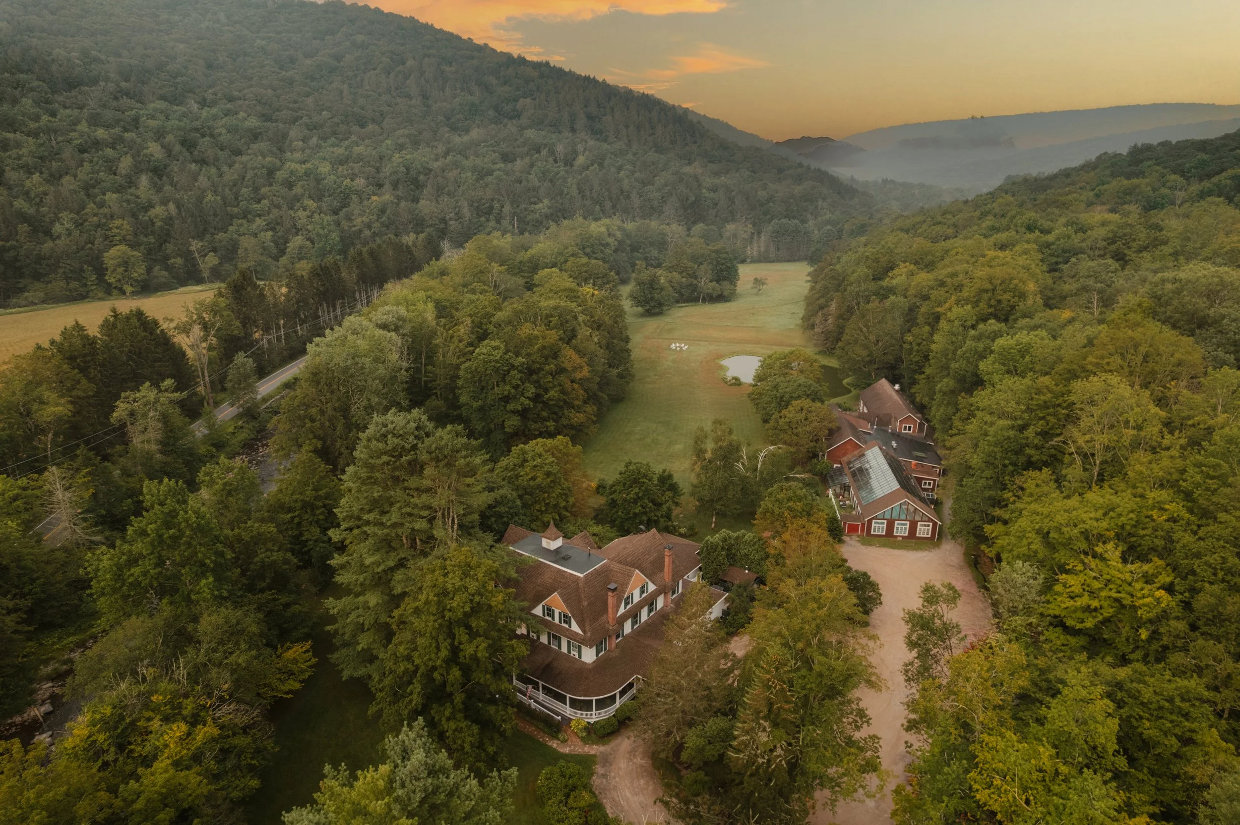 Aerial view of a large house and several outbuildings surrounded by dense green trees in a rural area with hills in the background, a small pond, and a sunset sky.