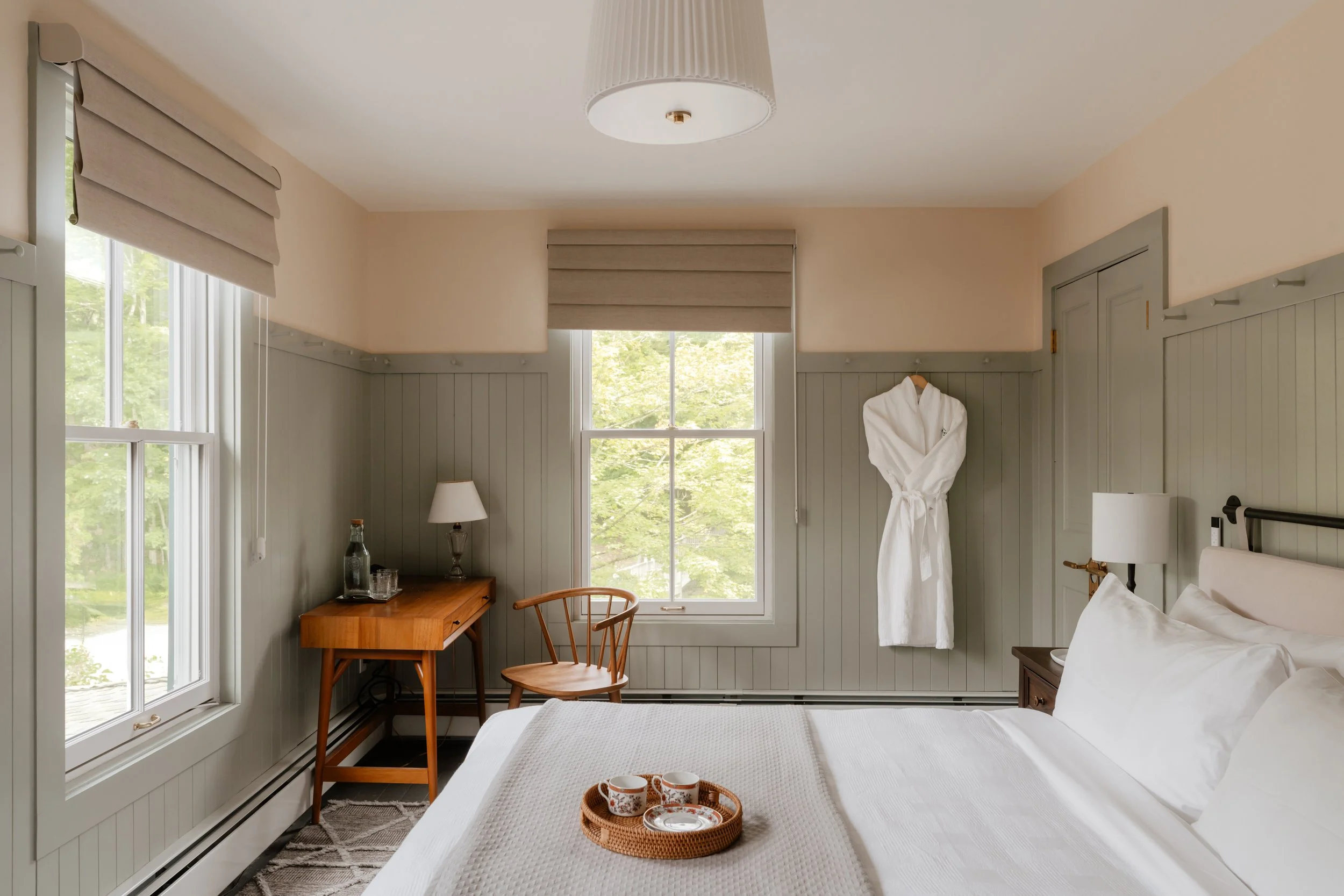A cozy bedroom with a bed, two windows, a small wooden desk with a lamp, a wooden chair, a white bathrobe hanging on the wall, and a tray with cups on the bed.