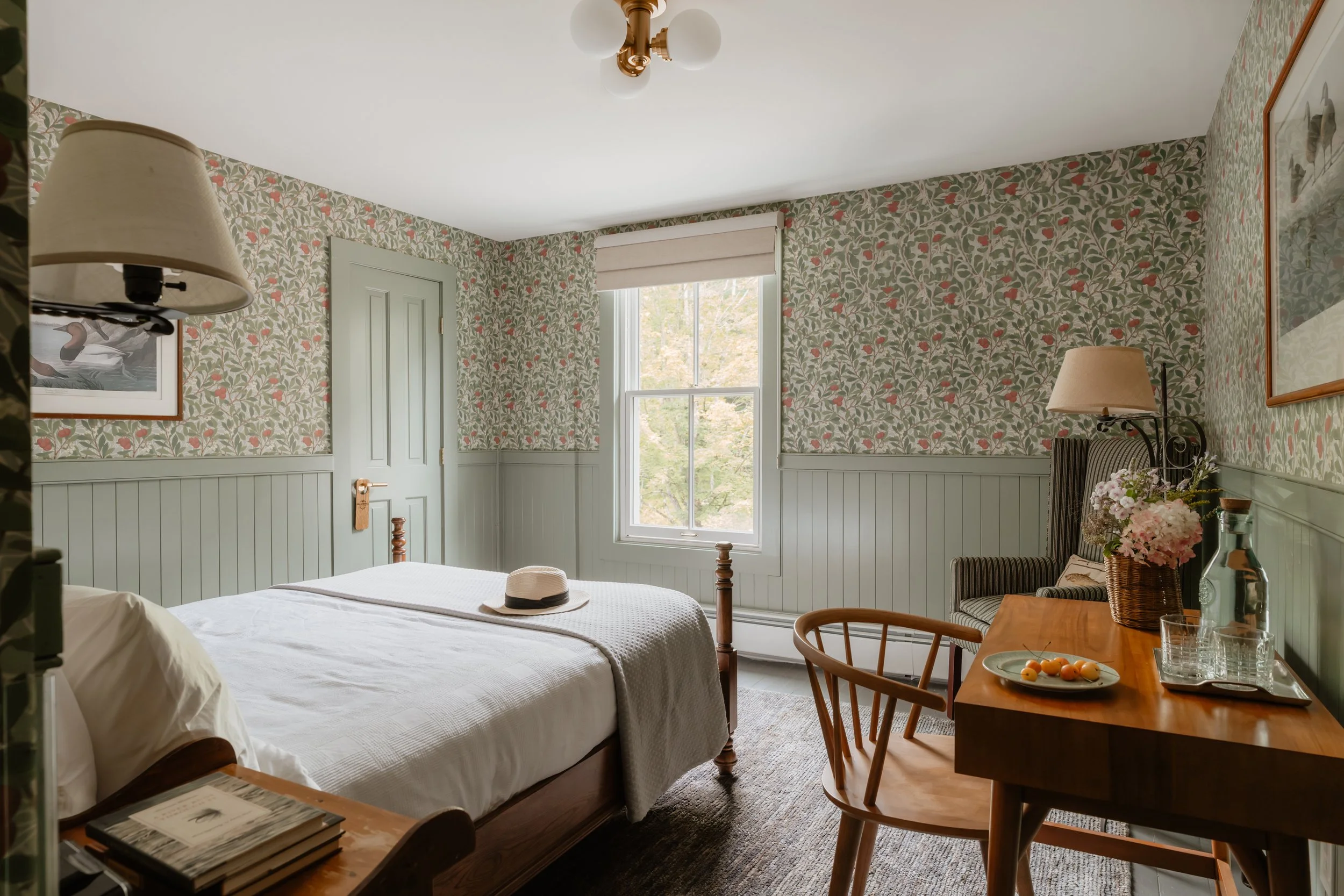 A cozy bedroom with floral wallpaper, a wooden bed with white bedding and a hat on top, a window with trees outside, a dresser with flowers, a chair, and a chair with black stripes, and framed art on the walls.