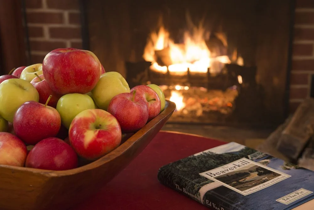 A wooden bowl filled with red and green apples on a table in front of a lit fireplace.