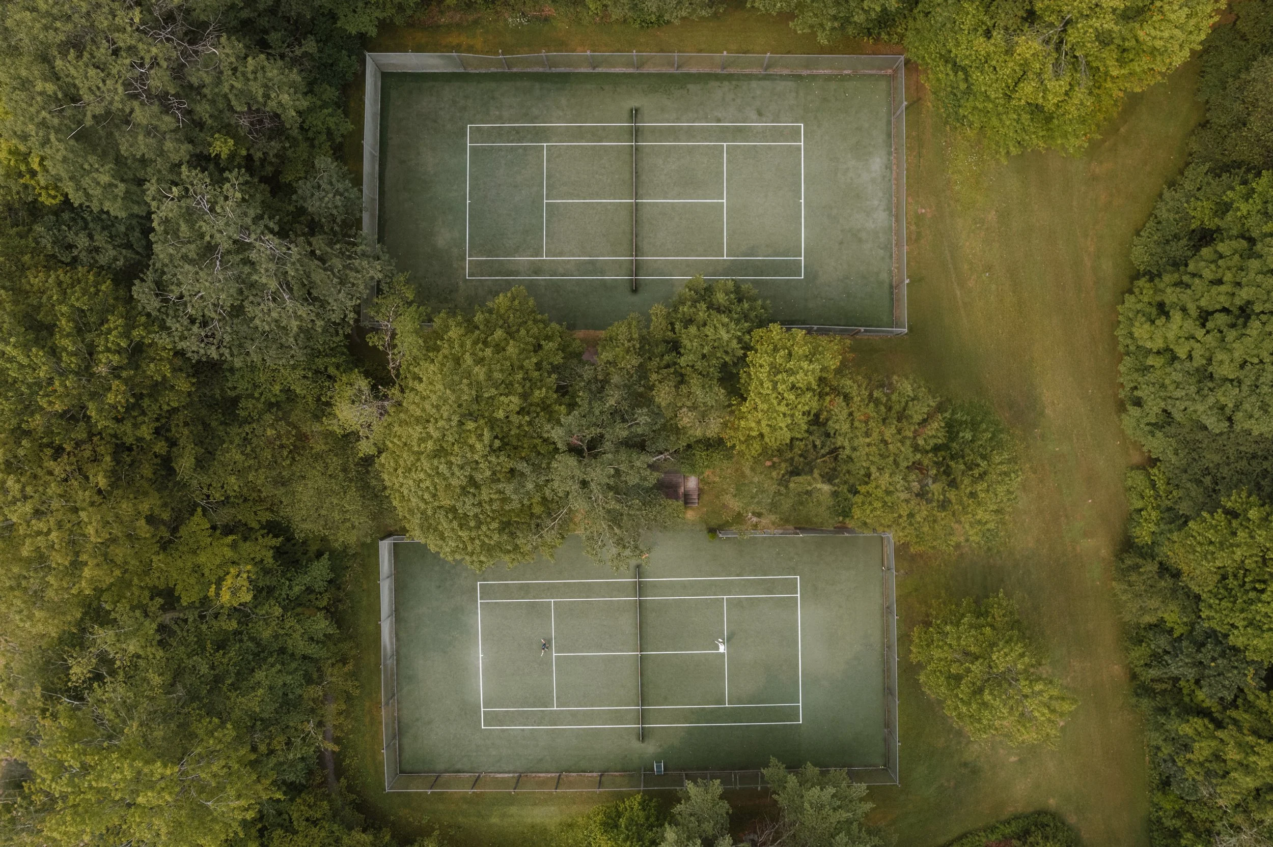 Aerial view of two tennis courts surrounded by trees, with a person visible on the lower court.