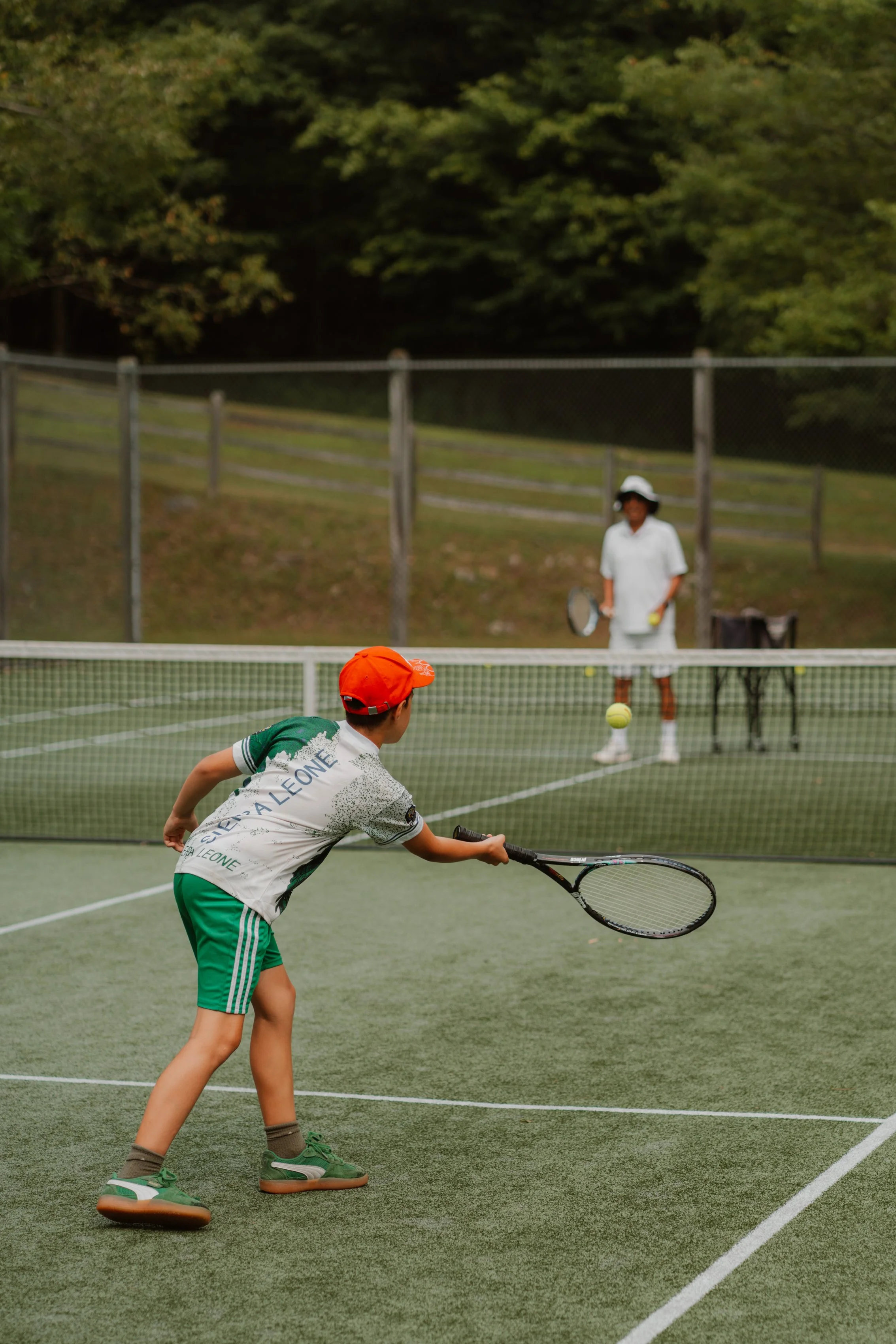 A young boy in a green and white sports outfit and orange cap playing tennis on an outdoor court, preparing to hit the tennis ball with a racket, while a girl in white sportswear and a white hat waits in the background.