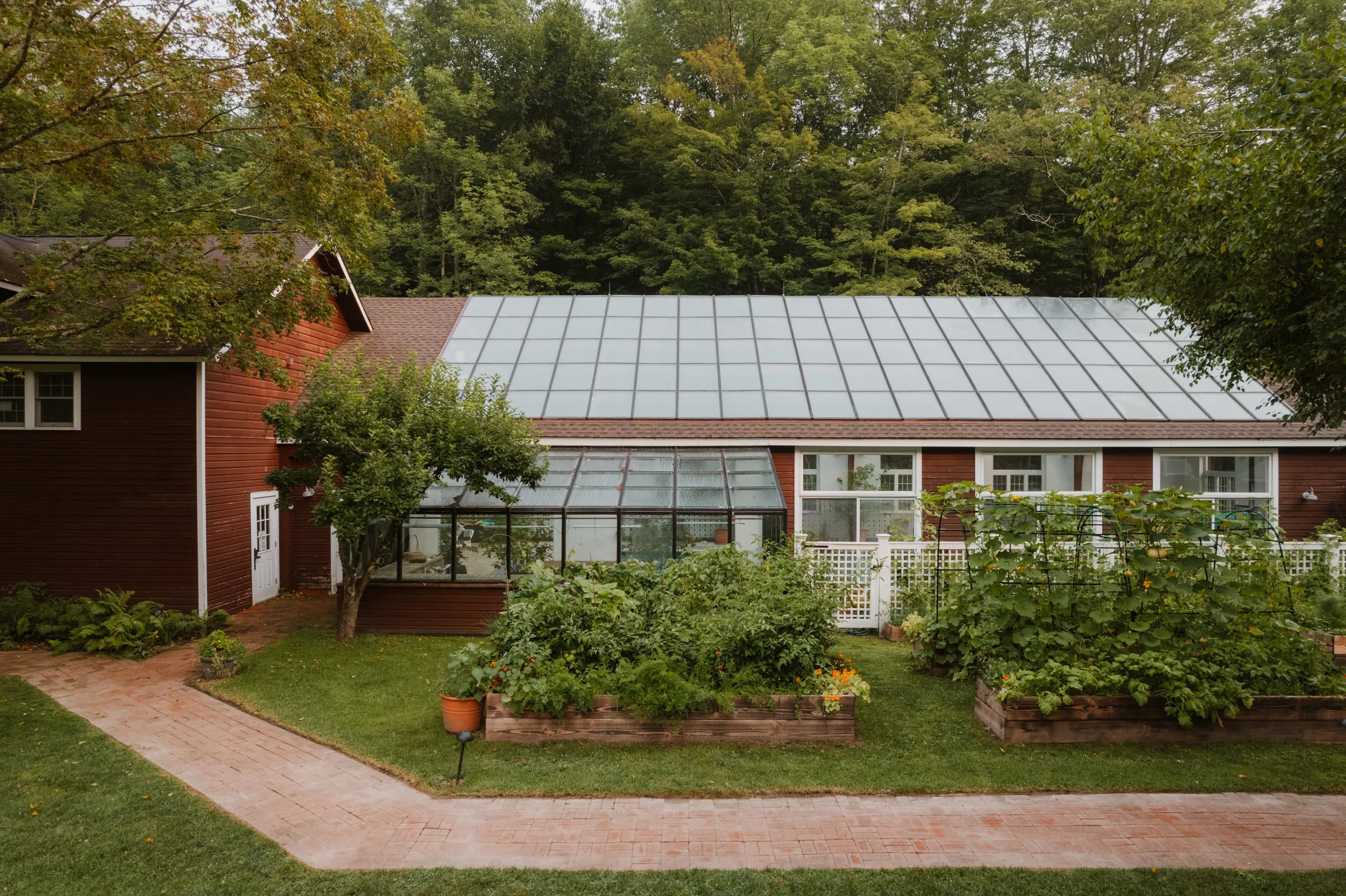 A house with a greenhouse on the back, surrounded by a garden with plants, trees, and a brick pathway, with a wooded forest in the background.