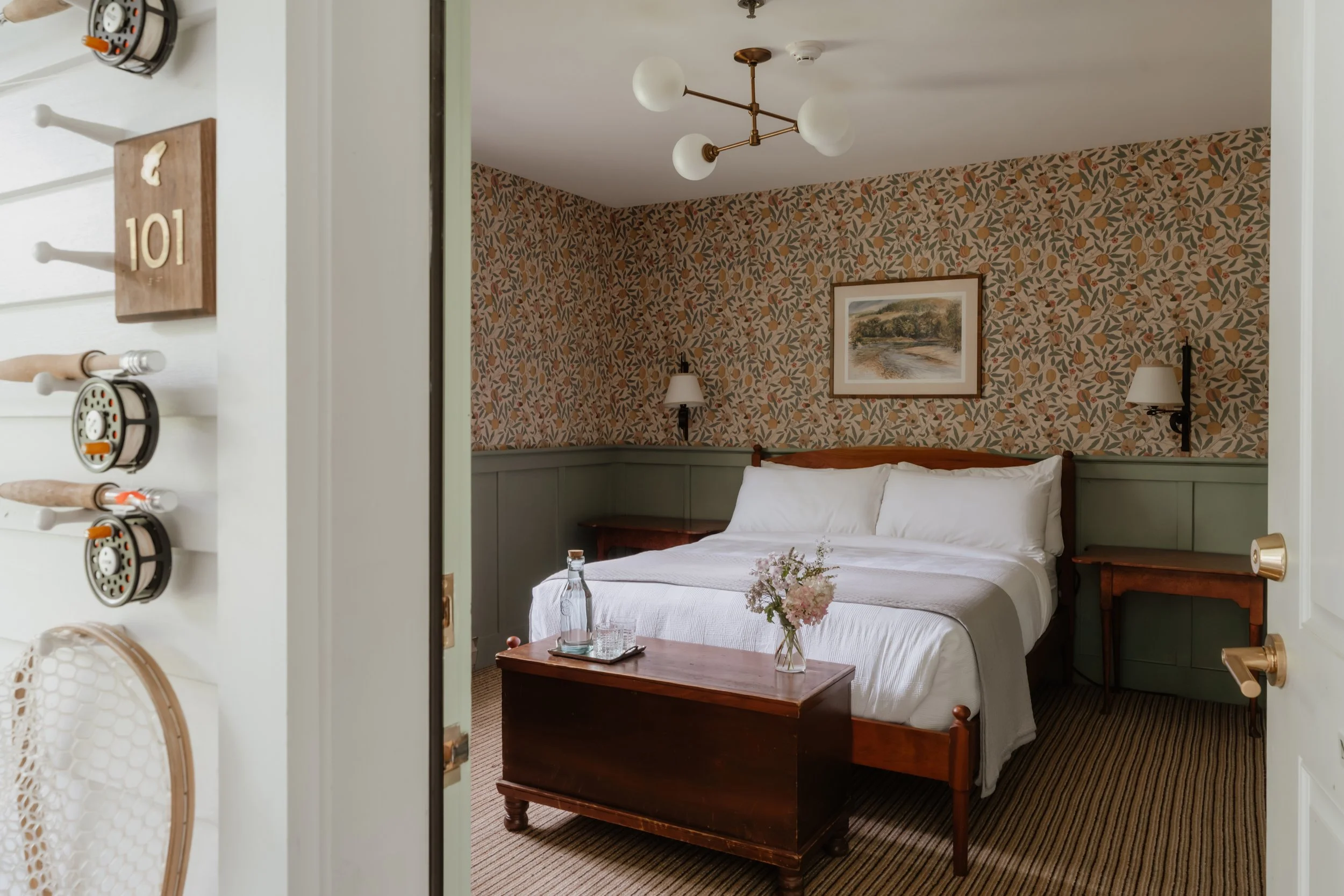 View of a cozy hotel bedroom with a bed, nightstands, wall sconces, and artwork, seen through an open door.