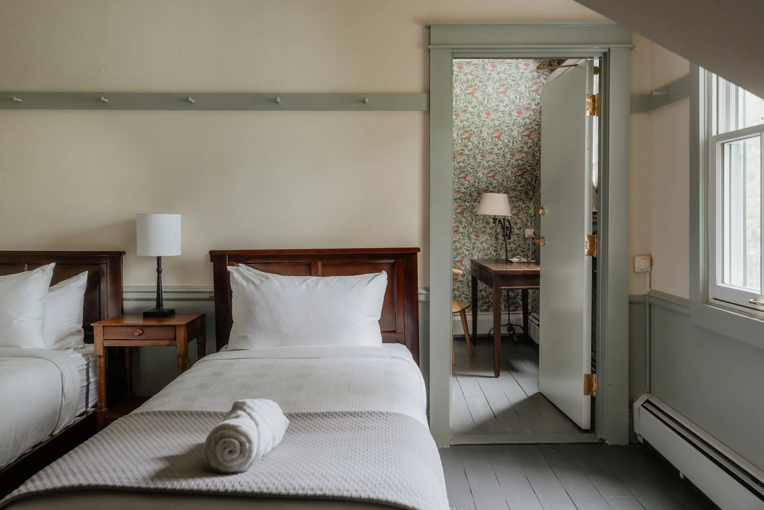 Hotel room with two beds, a nightstand with a lamp, and a small room with a desk, a lamp, and a chair visible through an open doorway.