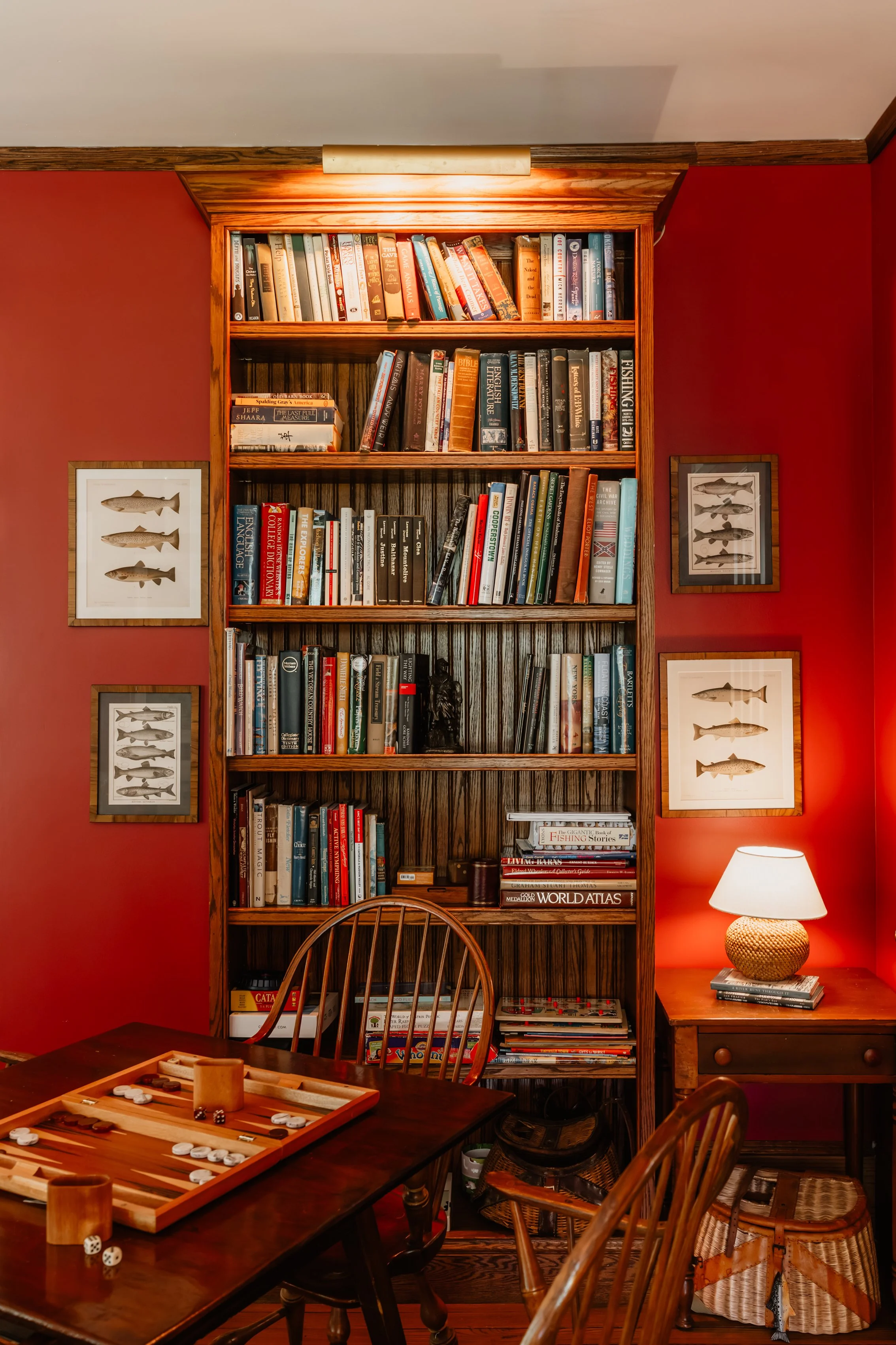A bookshelf filled with books against a red wall, with framed fish illustrations on either side, a wooden table with a backgammon game, and a table lamp on a small side table.
