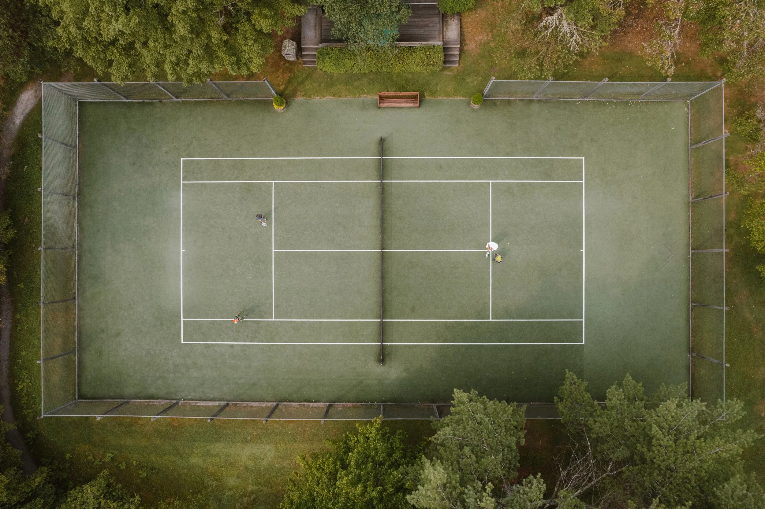 Aerial view of a tennis court with two players and a person sitting on the side with an umbrella, surrounded by trees.