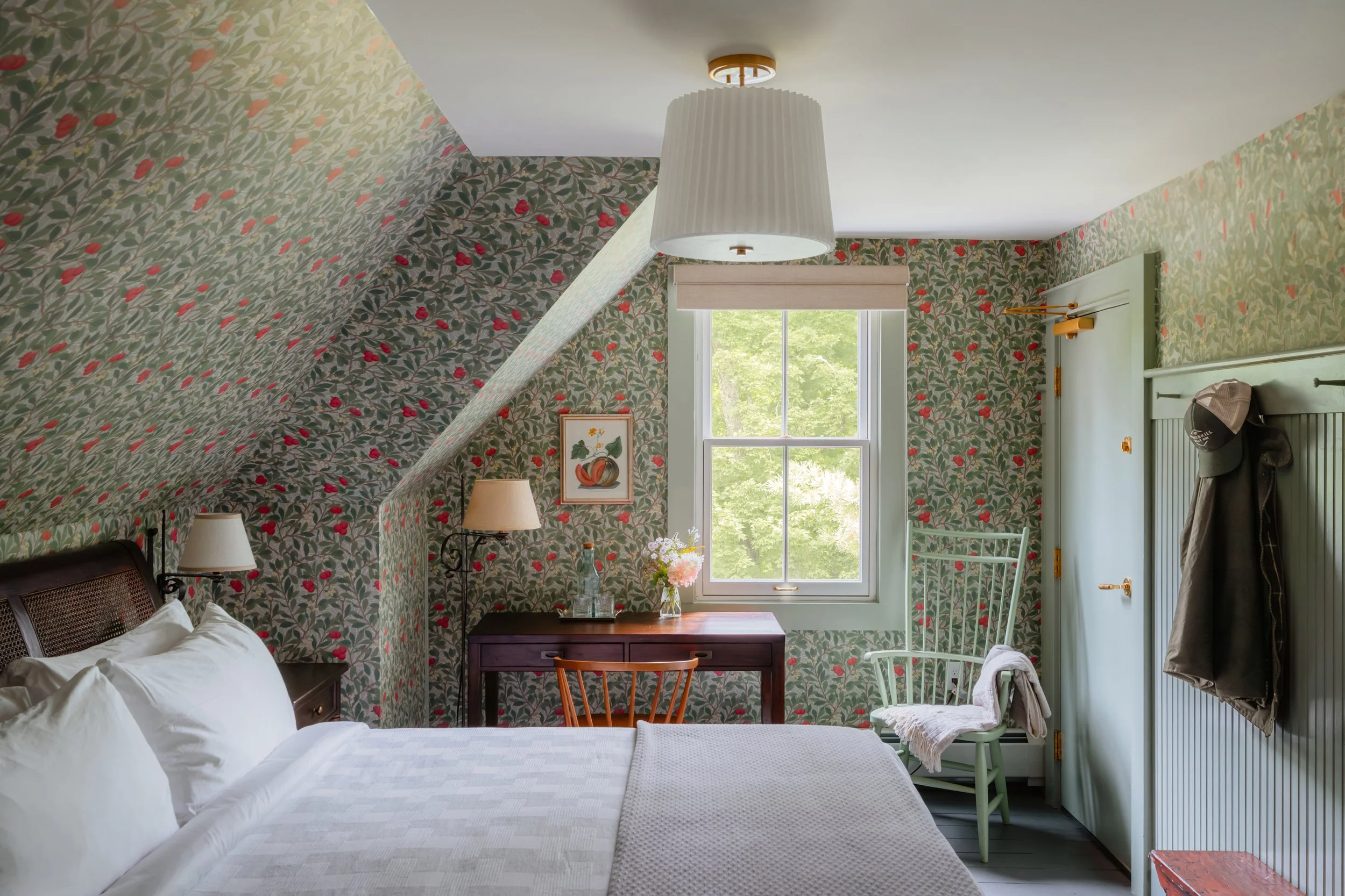 Cozy bedroom with floral wallpaper, bed with white linens, wooden desk near window, and a lavender chair with a blanket draped over it.