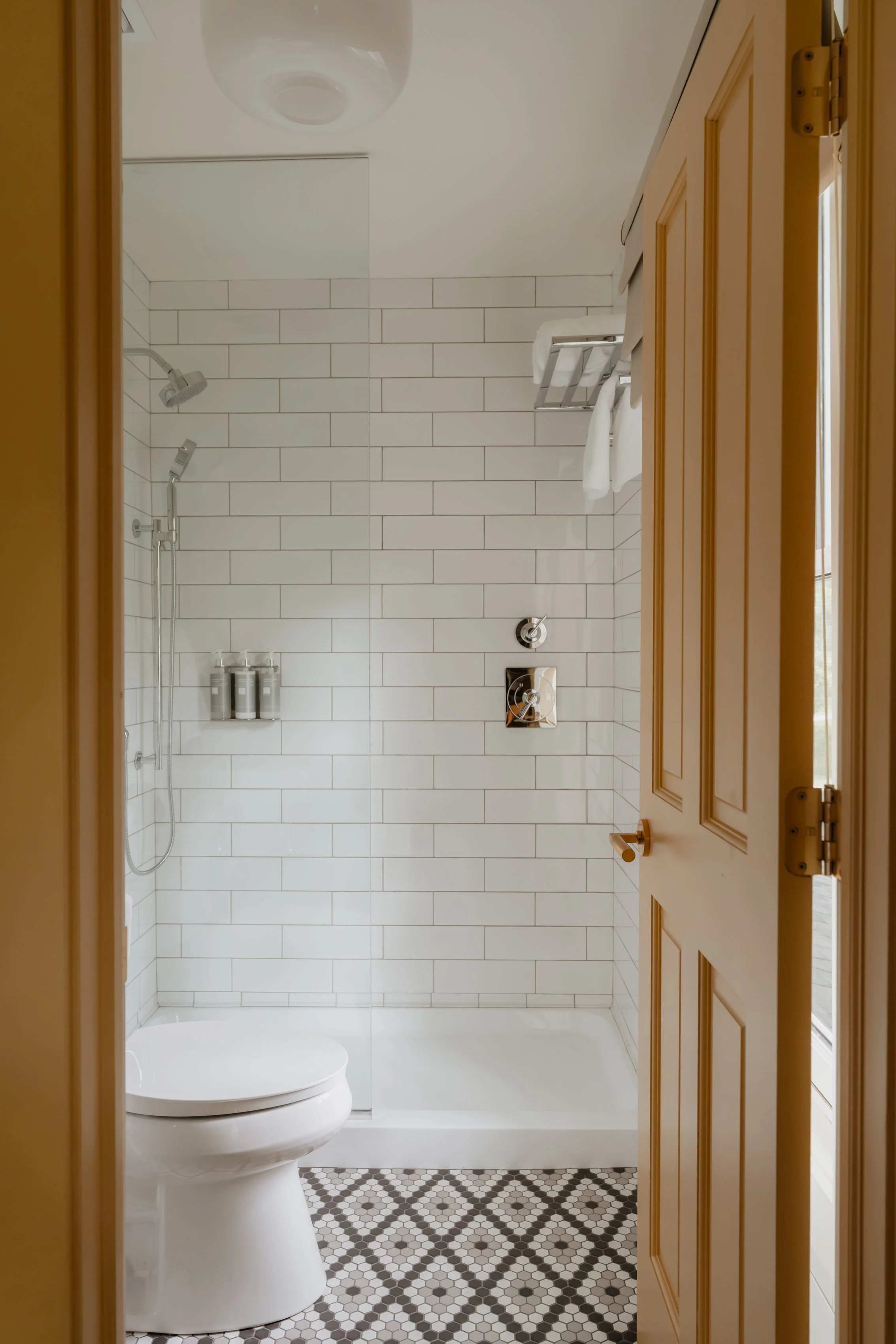 View of a modern bathroom with a white tiled walk-in shower, a toilet, and patterned floor tiles, seen through an open wooden door.