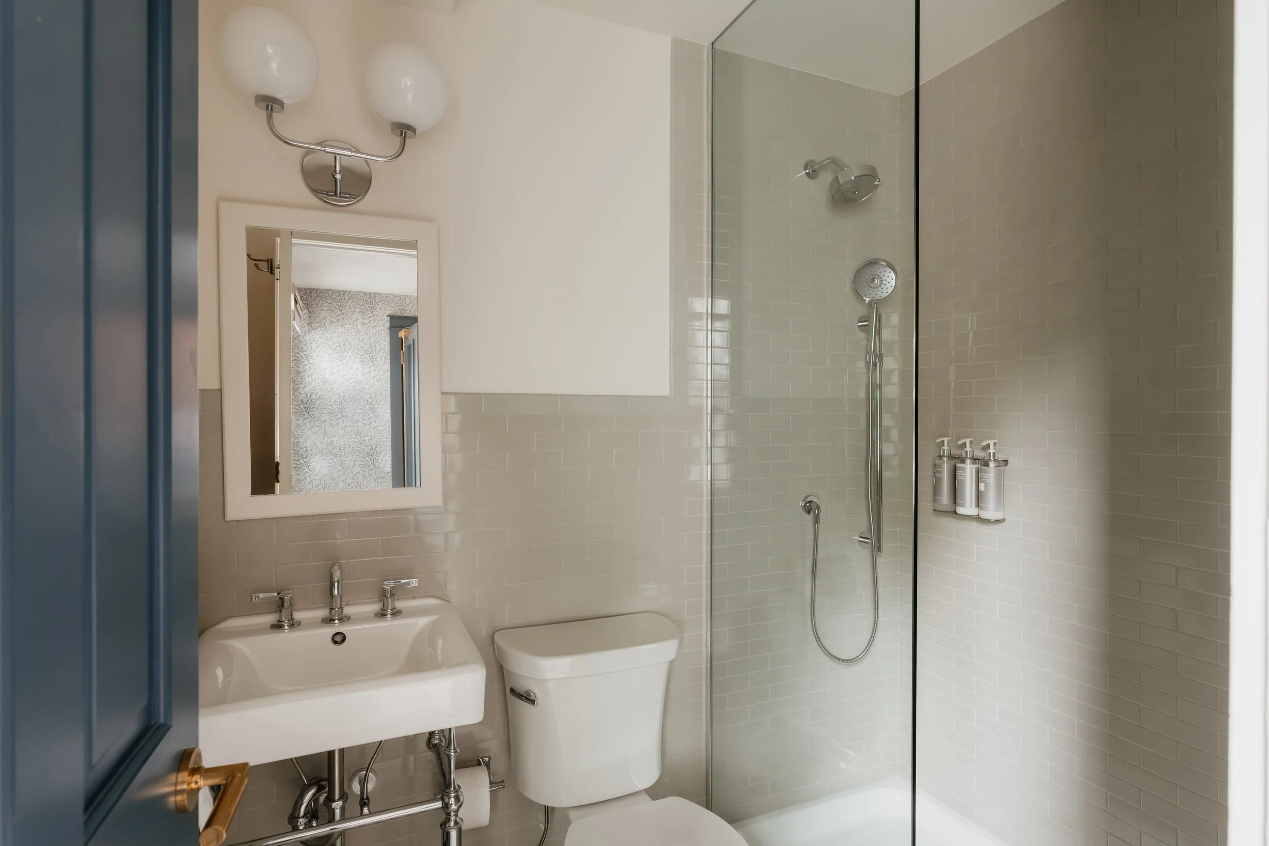 A modern bathroom with a white sink, mirror, toilet, and a glass-enclosed shower with beige subway tiles.