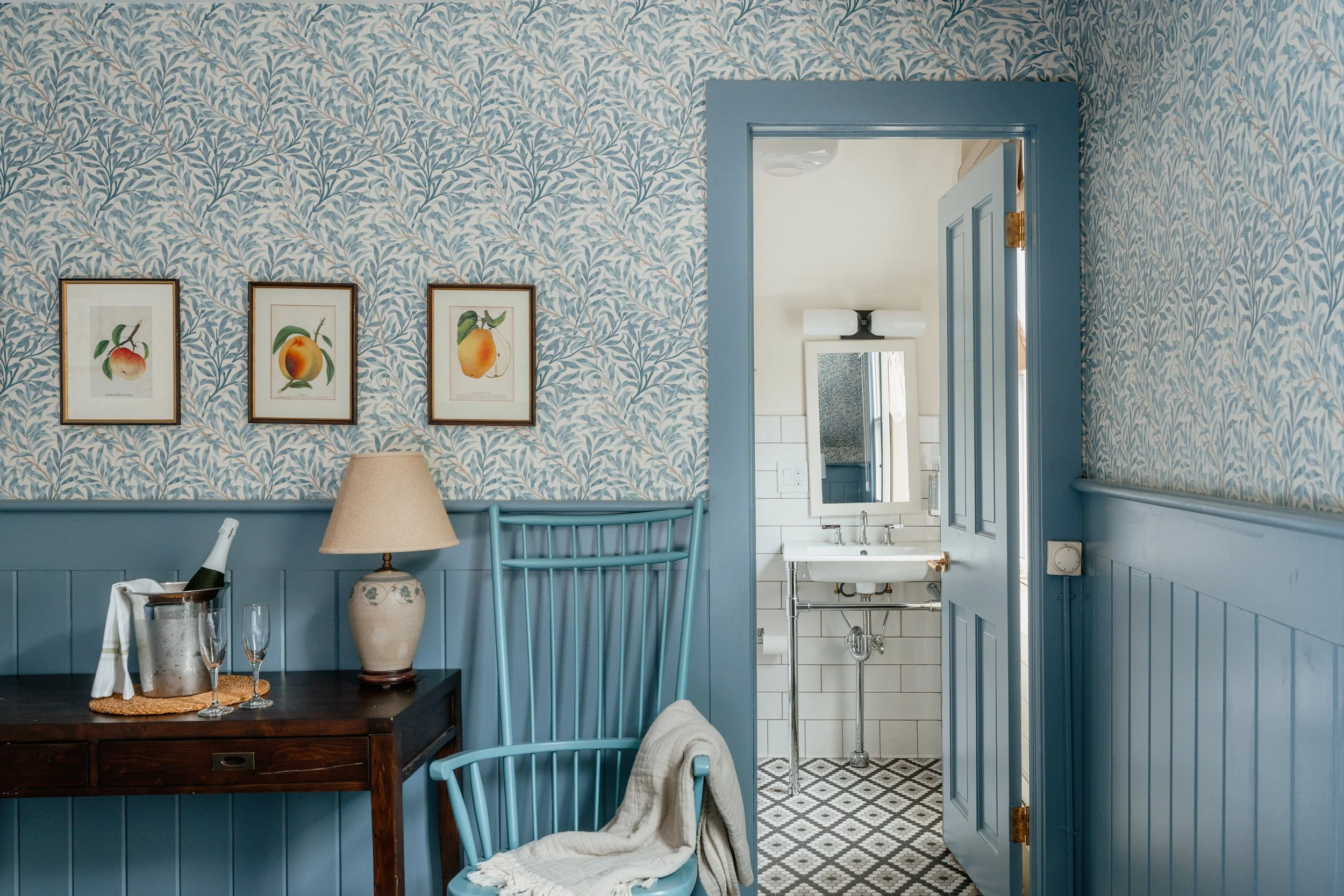 Interior of a room featuring patterned wallpaper with framed fruit botanical prints, a dark wooden table with a lamp, champagne bottle in an ice bucket, glasses, a blue wooden chair with a beige blanket, and an open door leading to a bathroom with wh