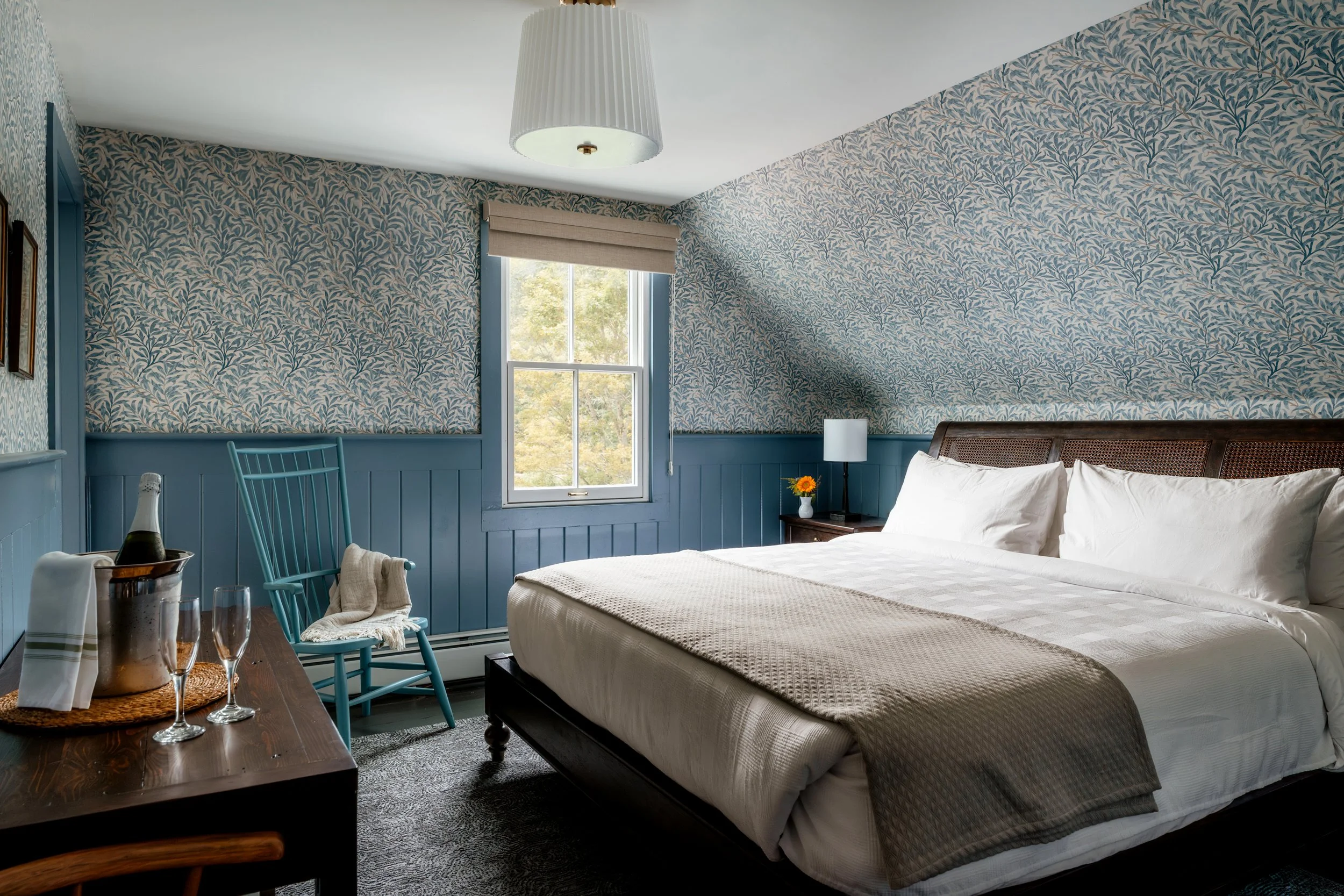 Cozy bedroom with a bed, nightstand with a lamp and vase, blue armchair, window with shades, patterned wallpaper, and a champagne bottle with glasses on a wooden table.