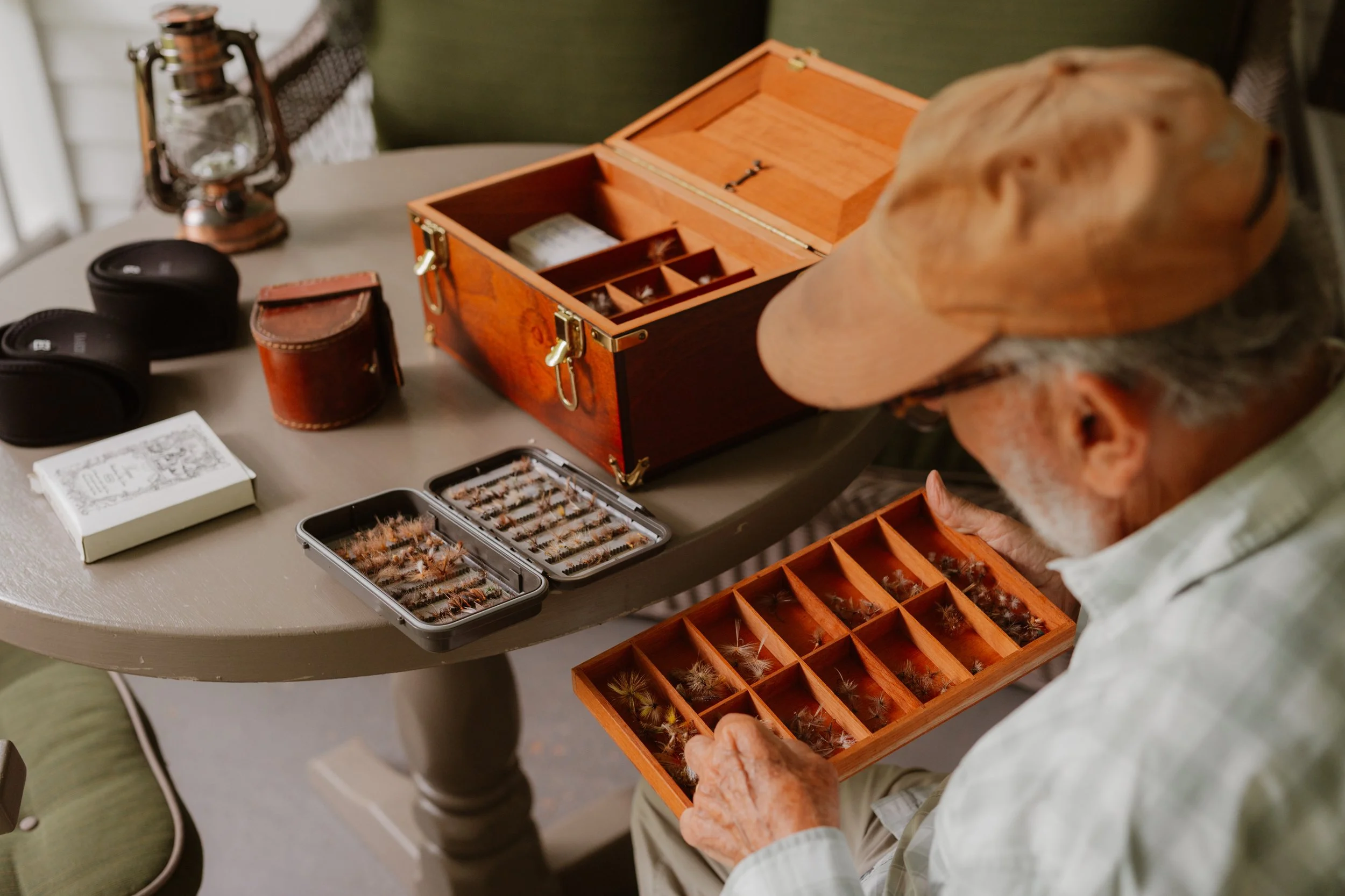 An elderly man arranging fly fishing lures in a wooden tackle box on a table with various fishing accessories, including binoculars and a lantern.