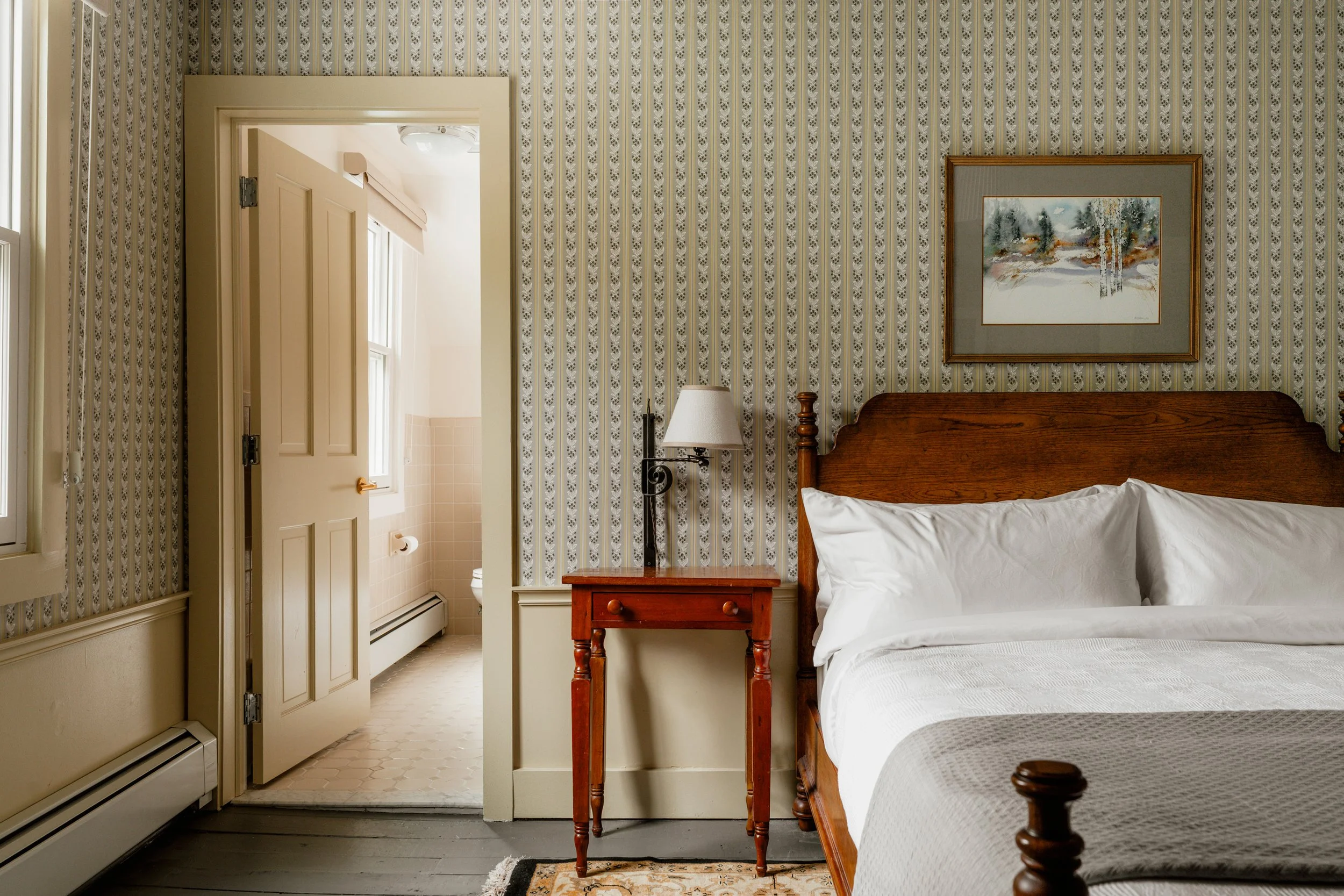 A cozy bedroom with vintage wallpaper, a wooden bed with white linens, a small wooden bedside table with a lamp, and a framed painting of a winter landscape, with an open door leading to a bathroom.