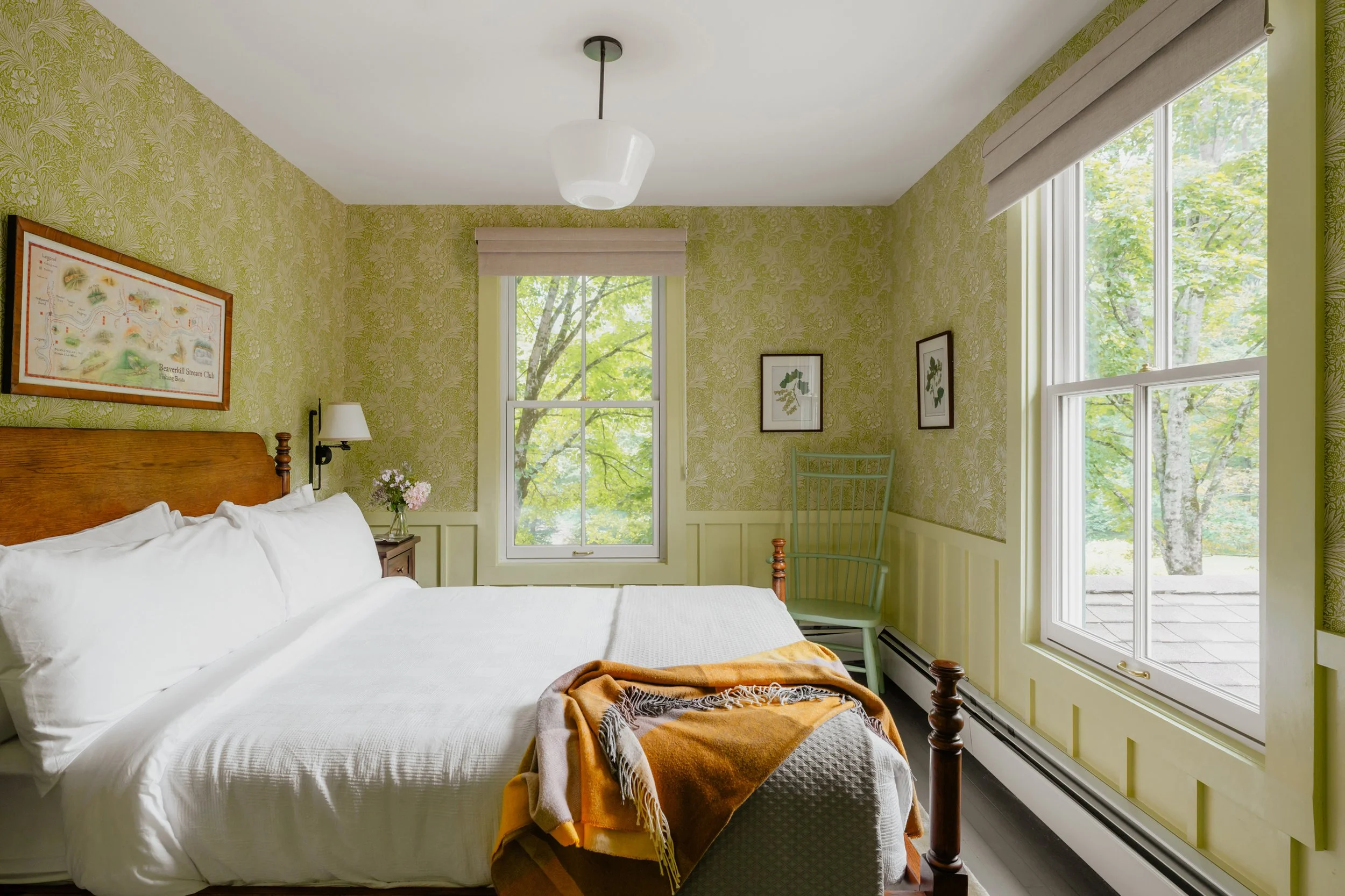 A cozy bedroom with yellow patterned wallpaper, a wooden bed with white bedding, a yellow and brown blanket, two windows with green views, a small green chair, framed wall art, a bedside table with flowers, and a ceiling light.