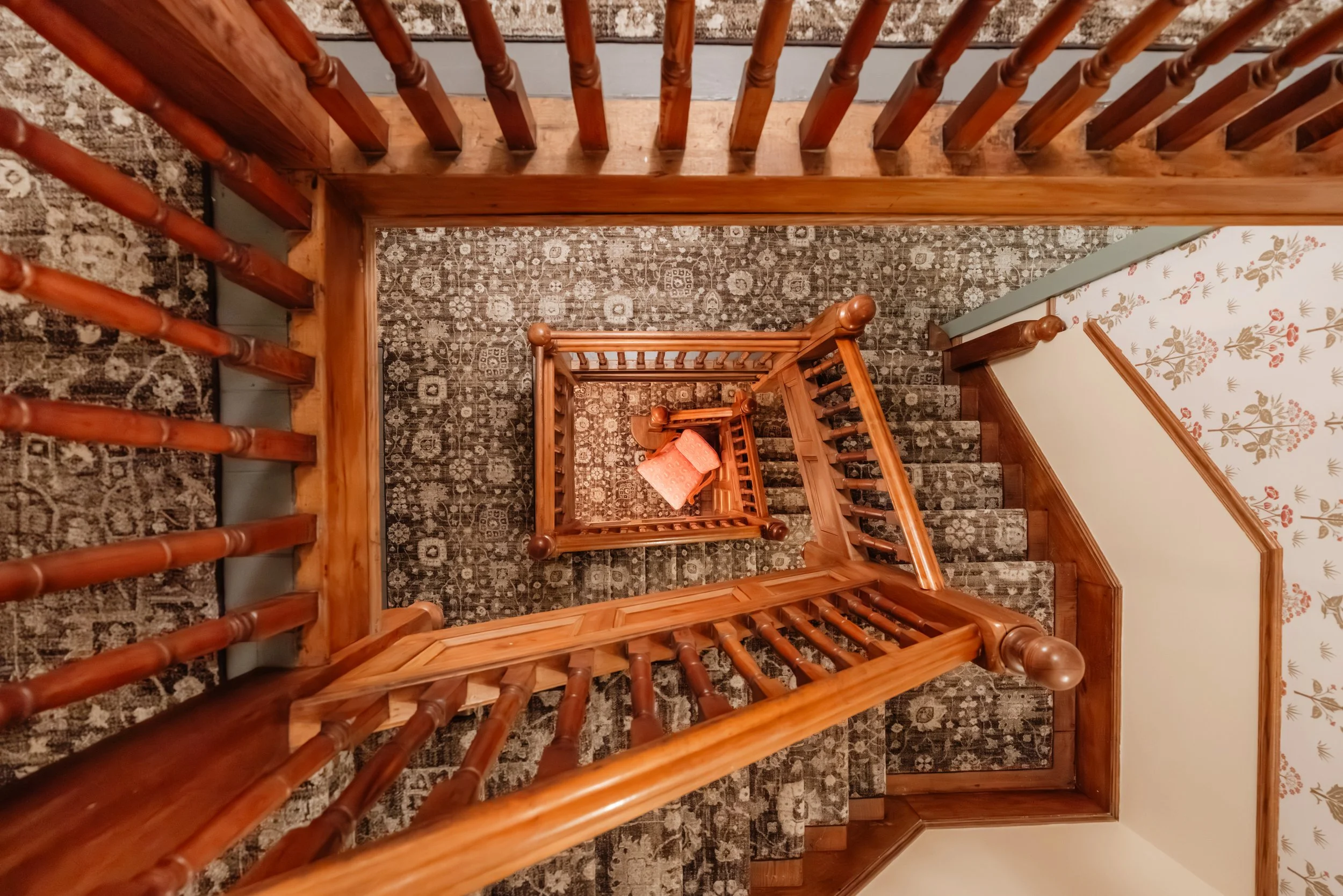 View looking down a wooden staircase with a patterned carpet runner and a small orange pillow at the bottom, with a floral wallpapered wall on the right.