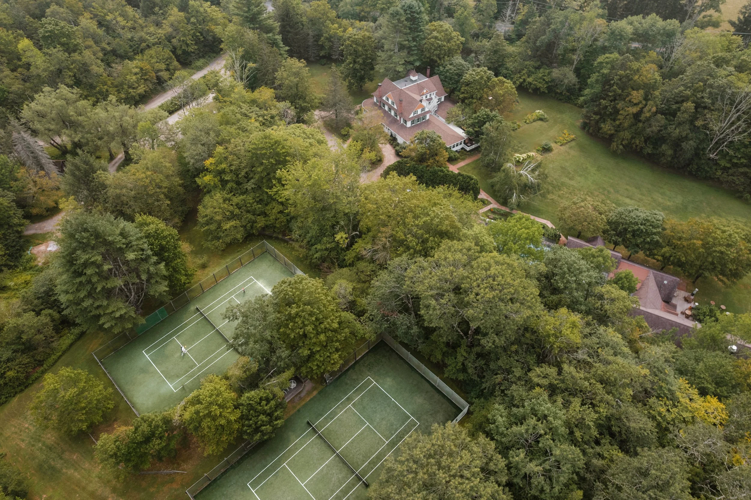 An aerial view of a large house surrounded by dense trees, with two fenced tennis courts and a smaller fenced paddle tennis court nearby.