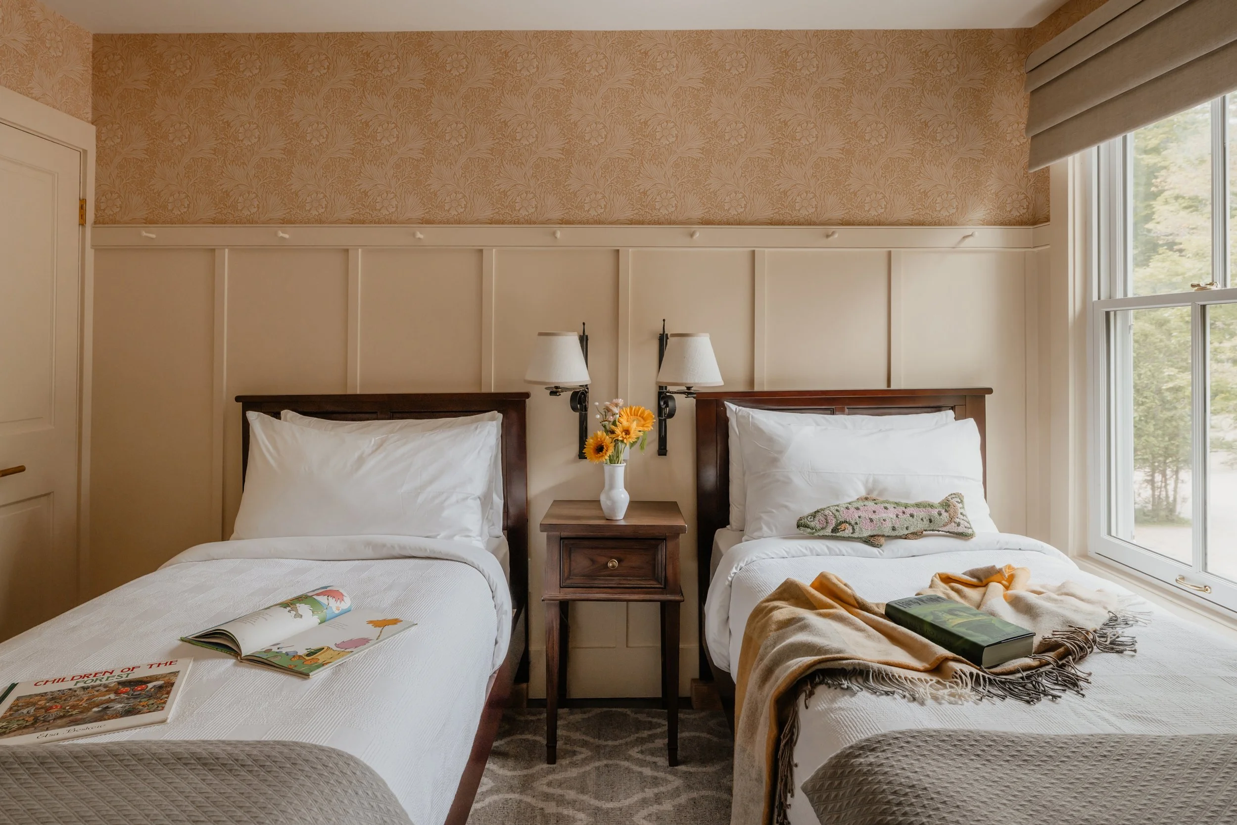 A bedroom with two twin beds, a wooden nightstand with a vase of sunflowers between them, wall-mounted lamps, a window on the right, and a patterned carpet.