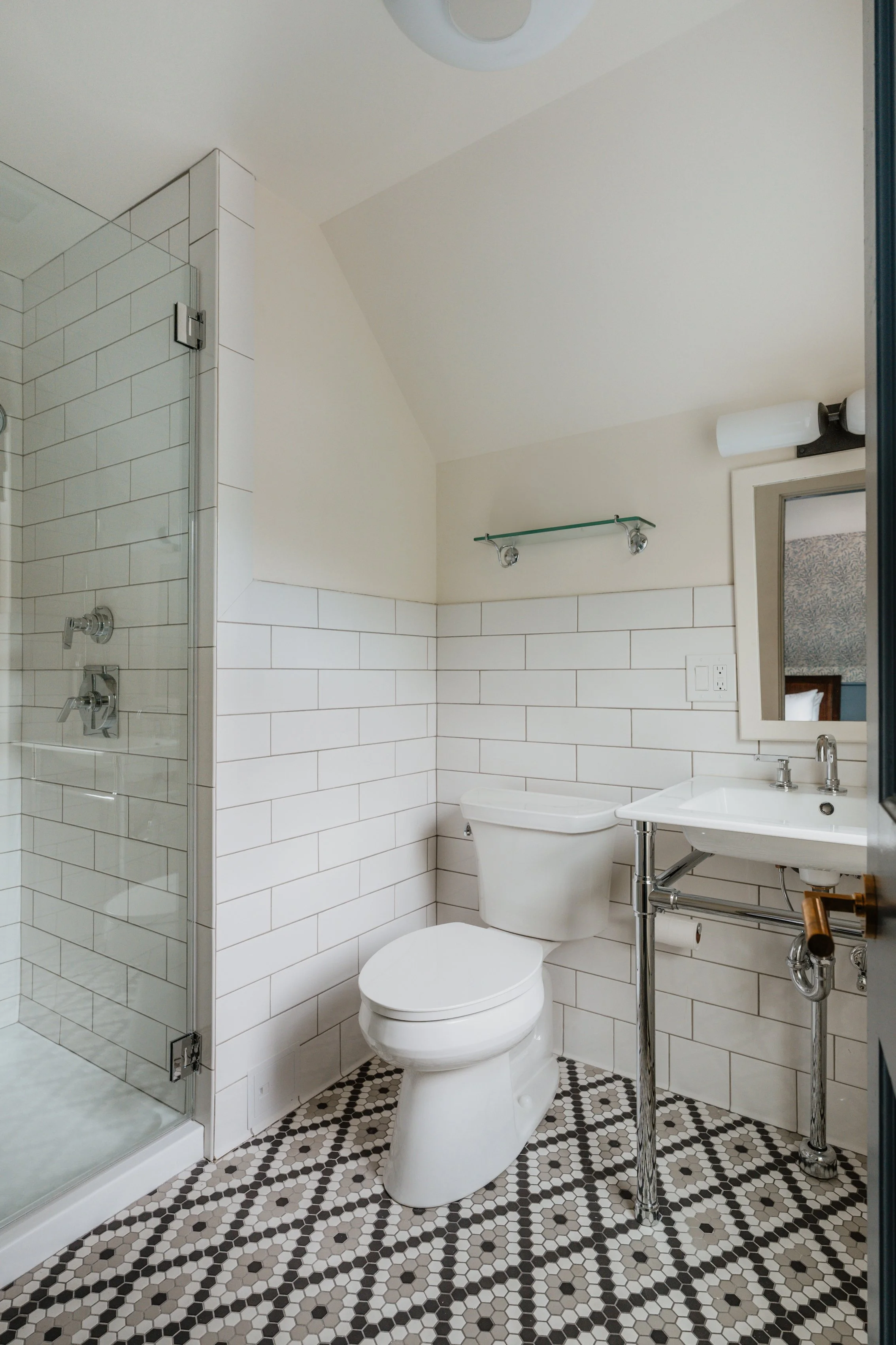Small bathroom featuring a shower with glass door, toilet, white sink with chrome pipes, mirror, and patterned tile floor.