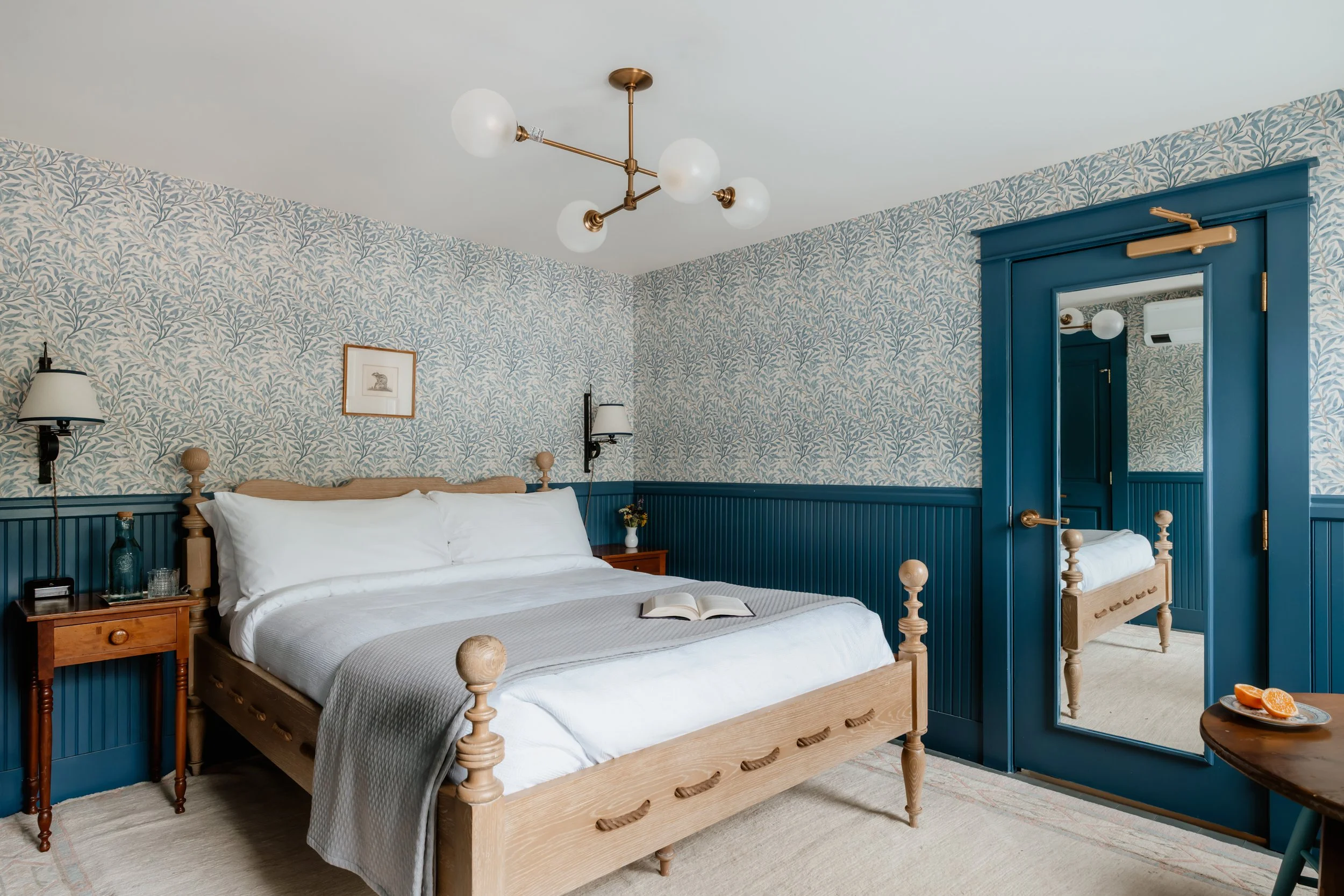 A bedroom with a wooden bed, white bedding, a blue wainscot, a blue door with a mirror, and a modern brass chandelier