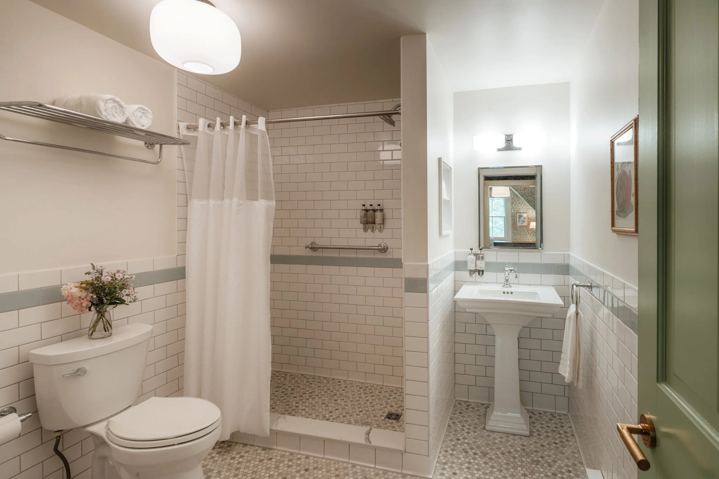 A clean, white-tiled bathroom featuring a bathtub with a shower curtain, a toilet with a flower vase on top, a pedestal sink with soap dispensers, a mirror, and a framed picture on the wall.